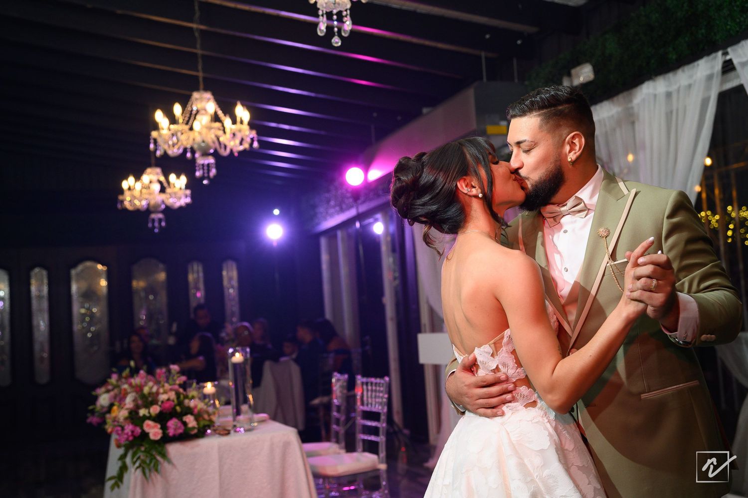 Bride and groom share a romantic kiss during their first dance at an elegant wedding reception with chandeliers and floral centerpieces.