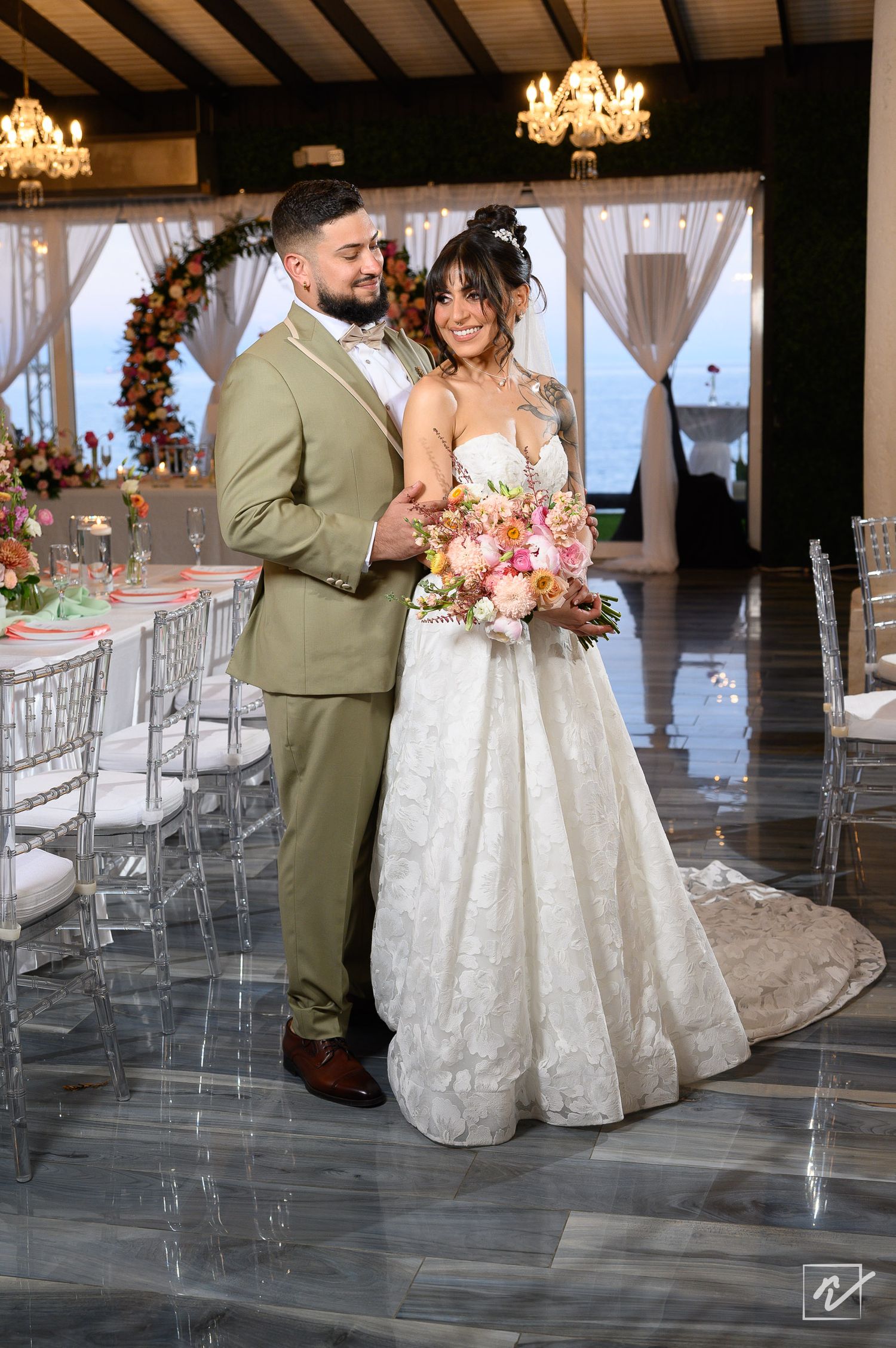 Bride in a lace gown holding a pink bouquet embraces a groom in an olive green suit in an elegant wedding venue with chandeliers.