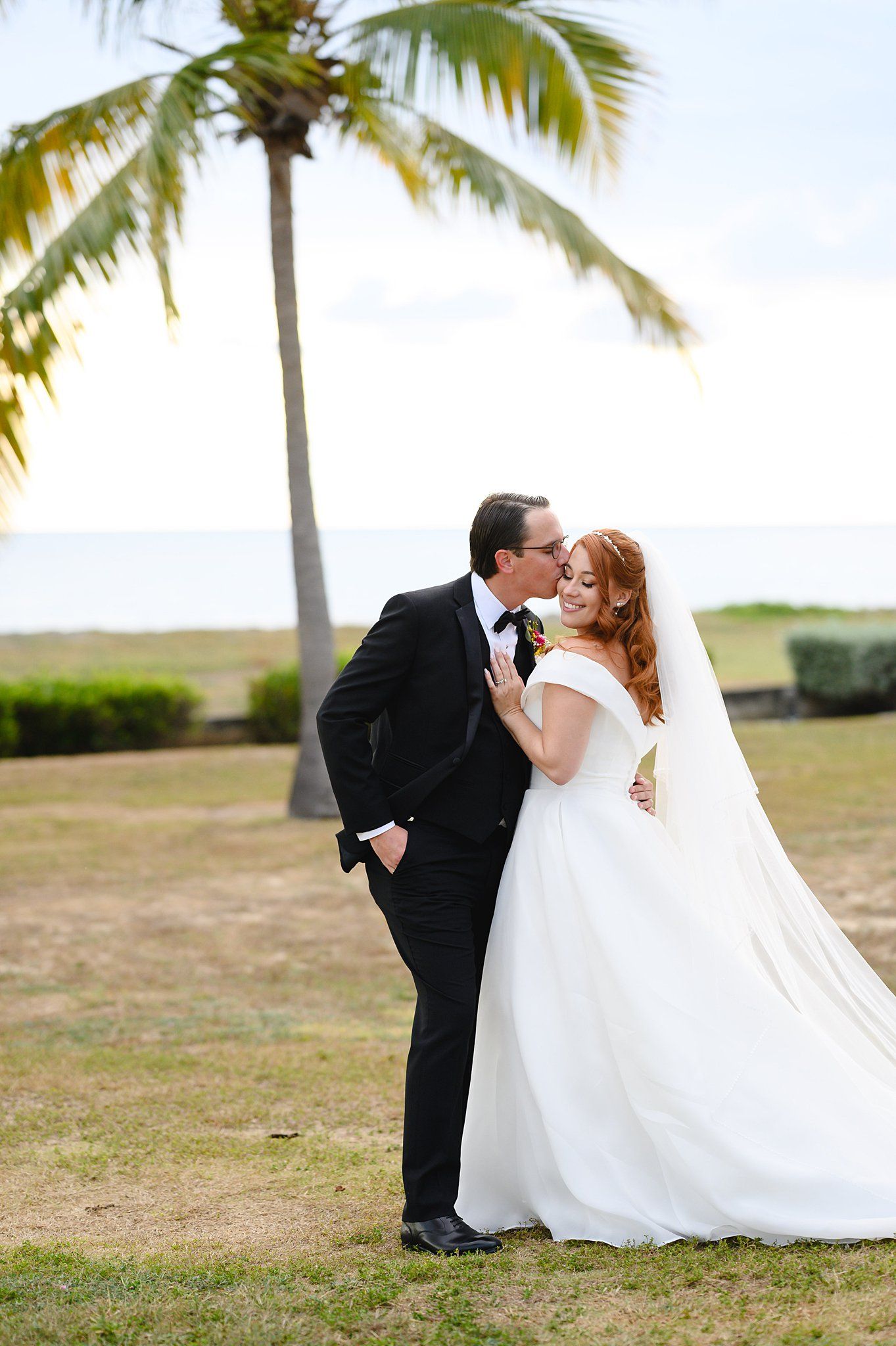 Bride and groom share a romantic kiss near a palm tree with ocean views at their tropical beach wedding.