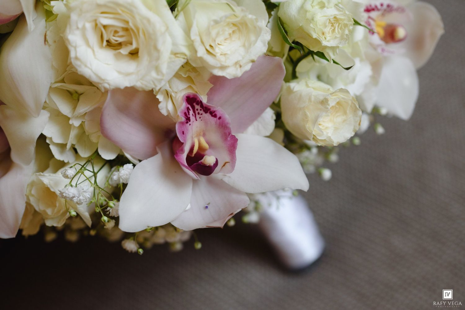 Close-up of a bridal bouquet featuring white roses, pink orchids, and delicate baby's breath on a gray surface.