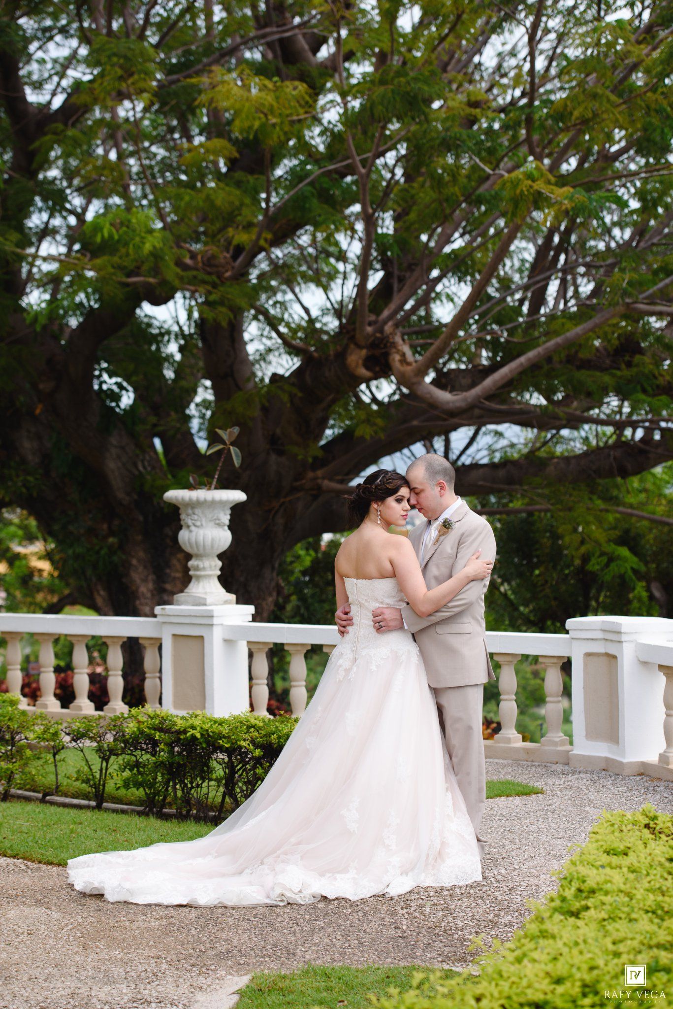 Bride and groom embracing on elegant white balustrade terrace surrounded by lush tropical gardens during outdoor wedding ceremony.