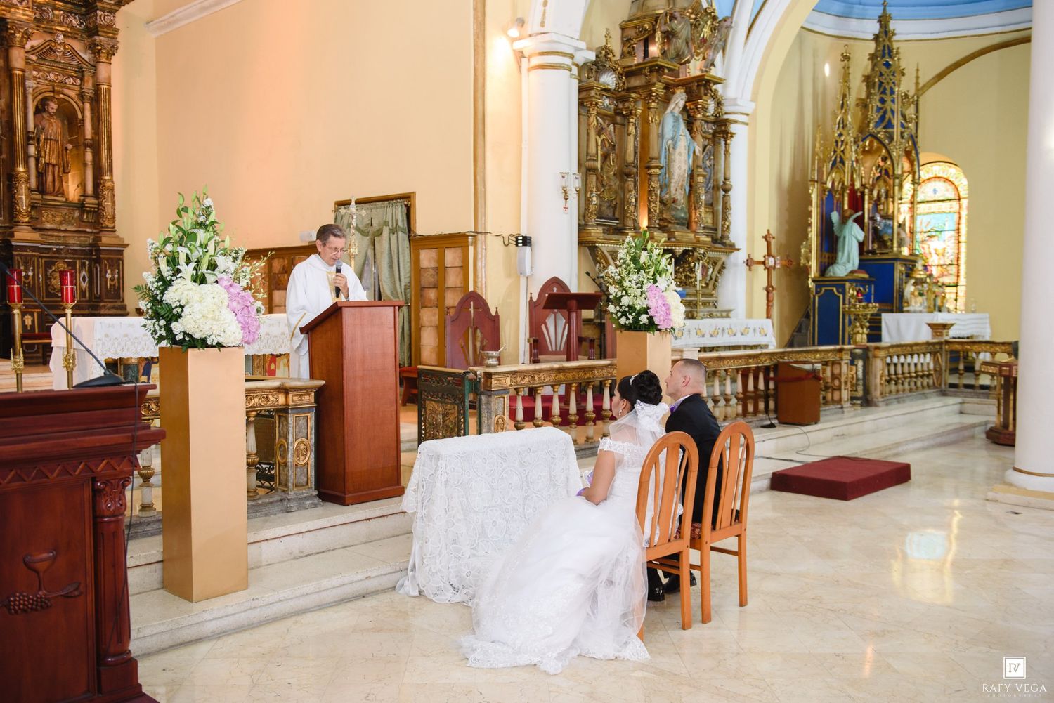 Bride and groom kneel at altar during Catholic wedding ceremony in ornate church with floral decorations.
