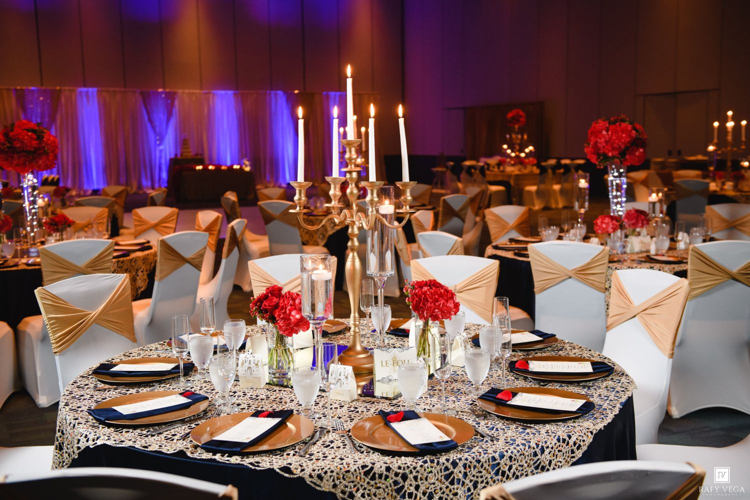 Elegant wedding reception hall with gold candelabras, red floral centerpieces, black lace tablecloths, and gold sash chairs.