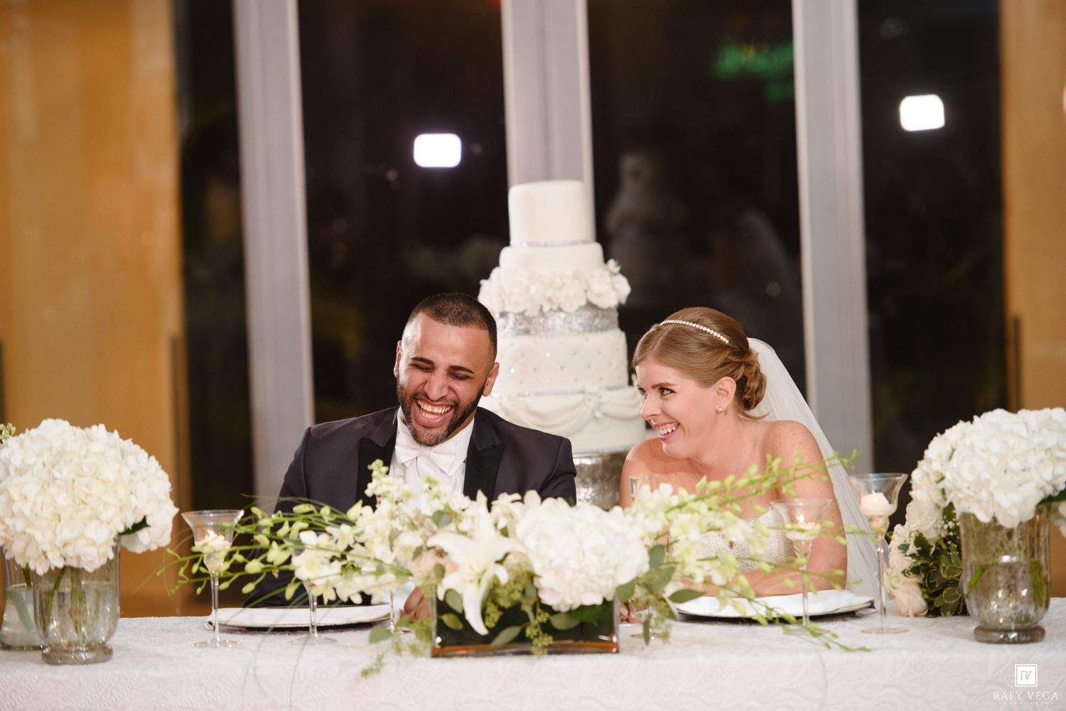 Bride and groom laughing at sweetheart table with white floral arrangements and tiered wedding cake in background.