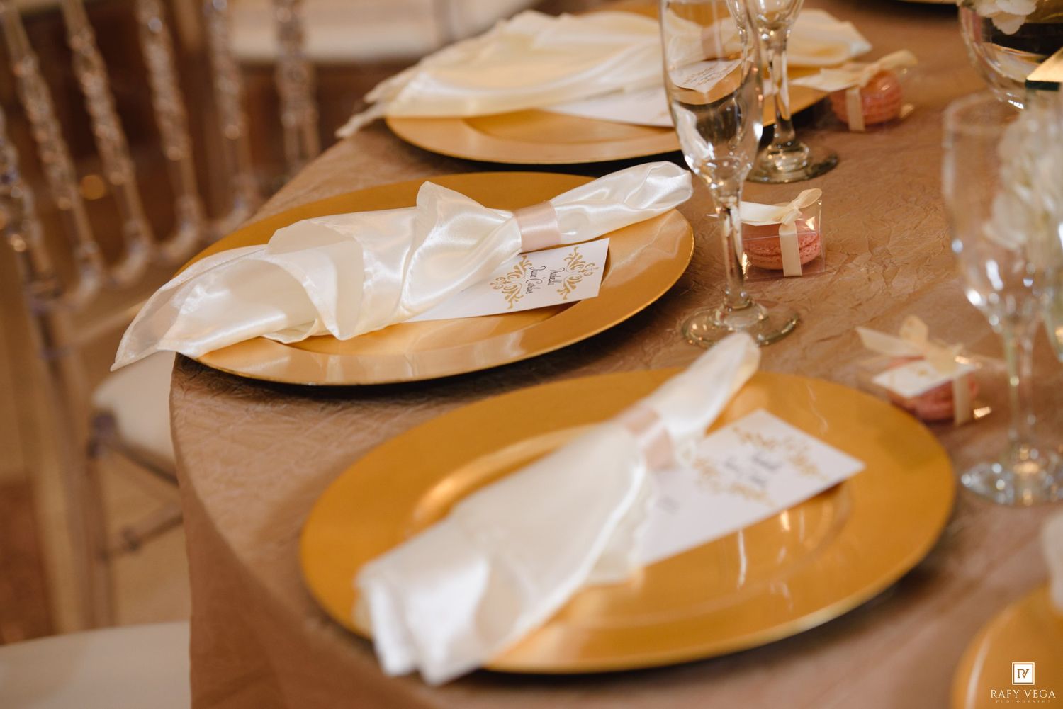 Elegant gold charger plates with white folded napkins and place cards on a champagne tablecloth at a wedding reception.