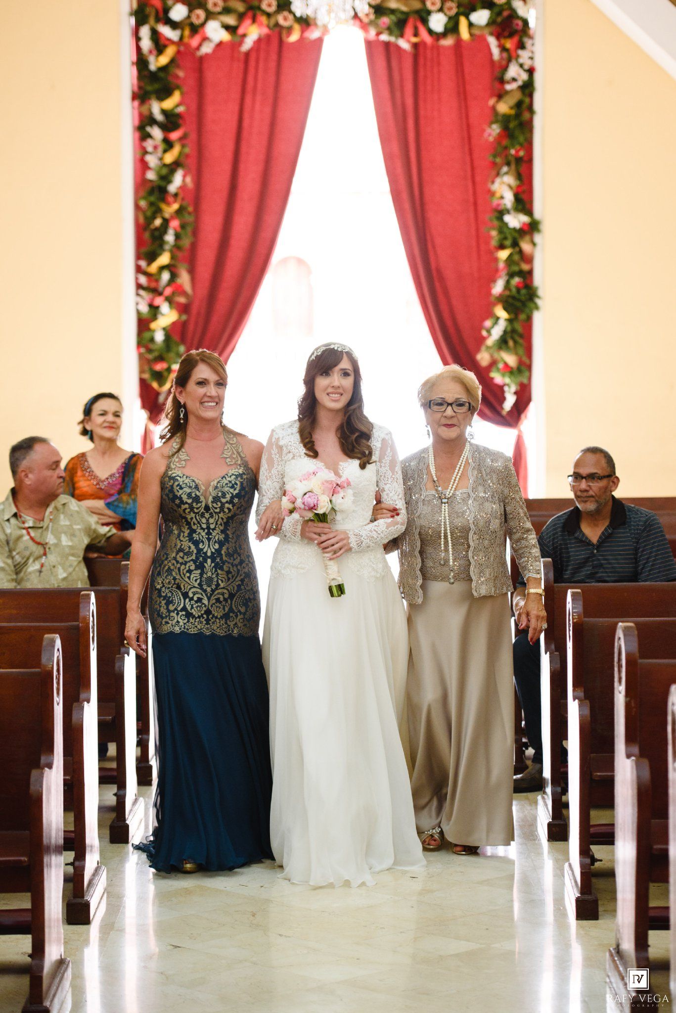 Bride walks down church aisle in white gown with bouquet, flanked by two women in formal attire.