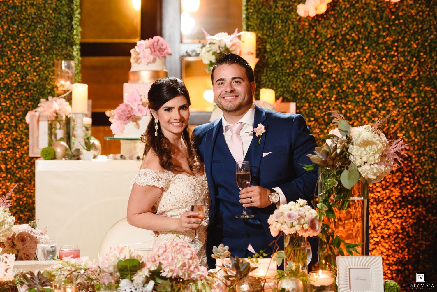 Bride and groom smiling at wedding reception table with pink floral arrangements and champagne glasses.