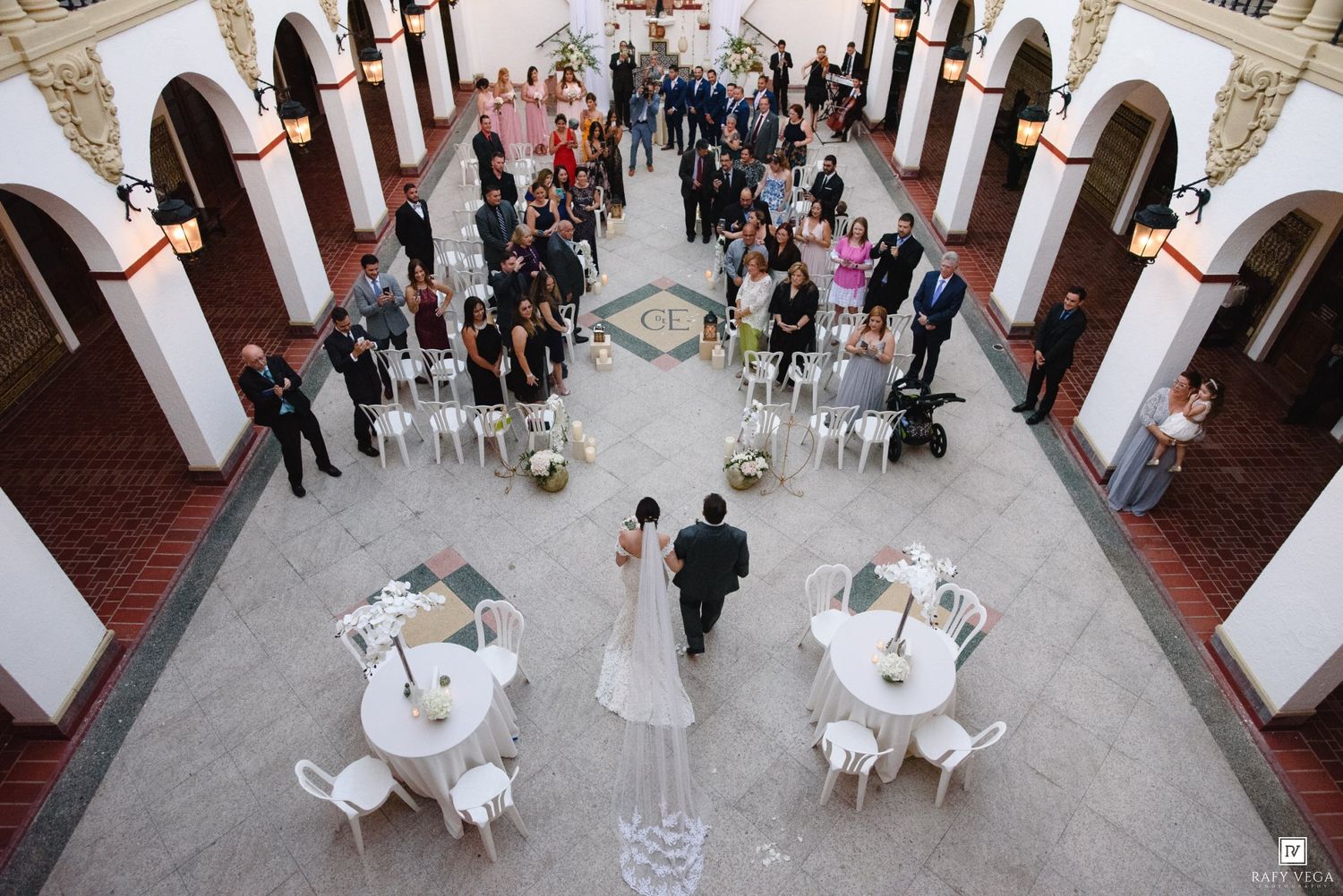 Aerial view of a wedding ceremony in an elegant courtyard with guests seated in white chairs around the bride and groom.