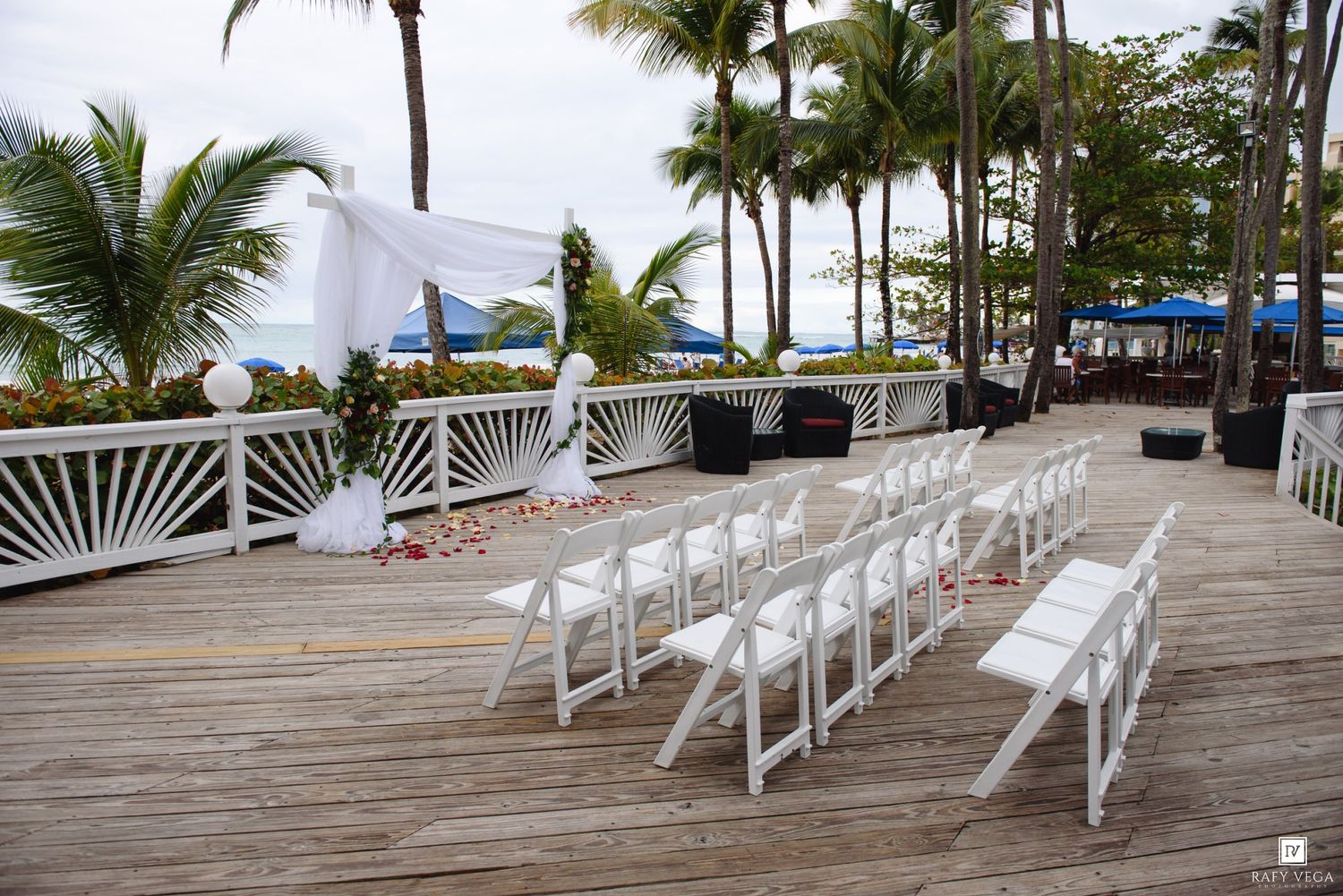 Outdoor beachside wedding ceremony setup with white chairs and floral arch on a wooden deck surrounded by palm trees.