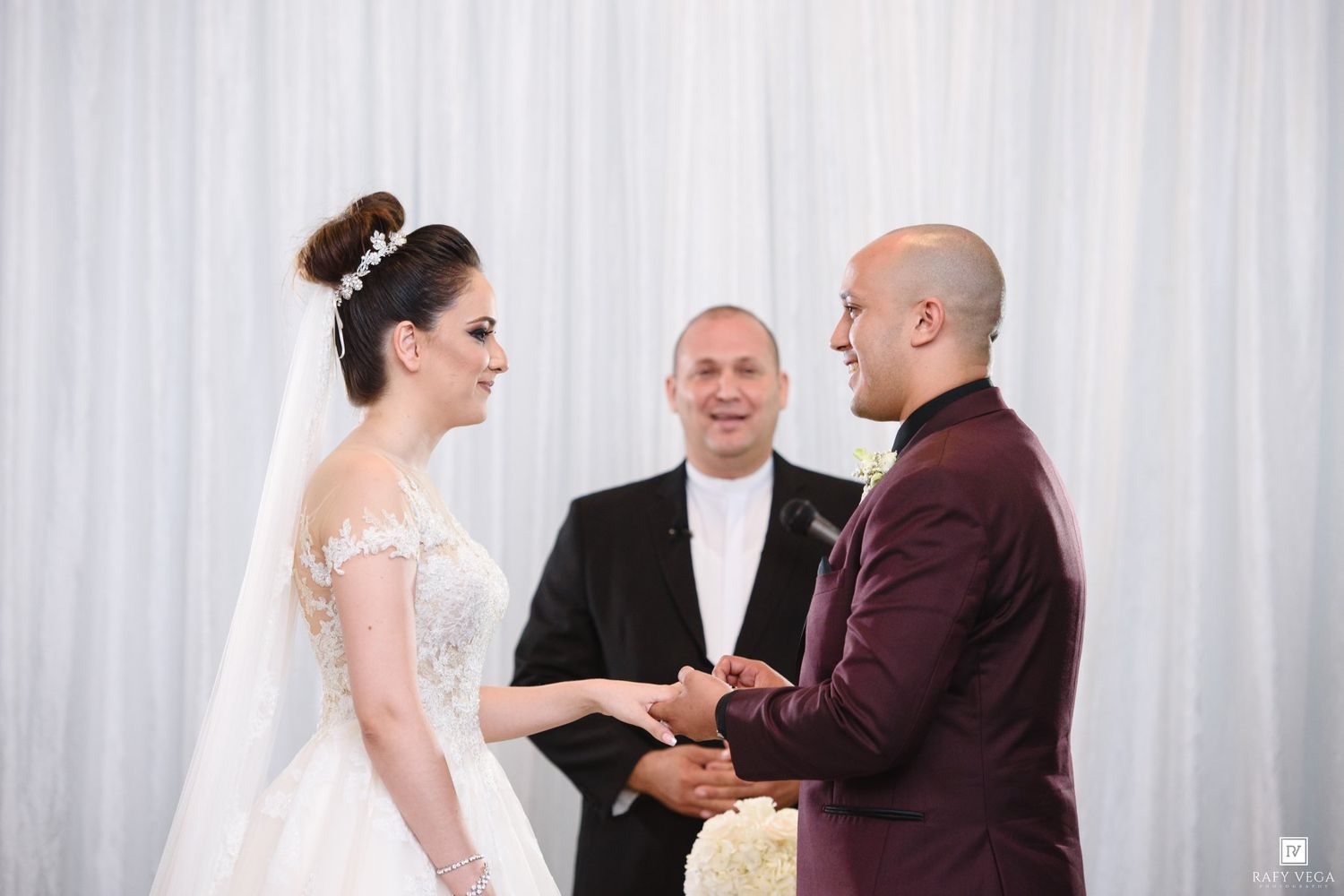 Bride and groom exchange vows at wedding altar while officiant presides over elegant ceremony with white drape backdrop.