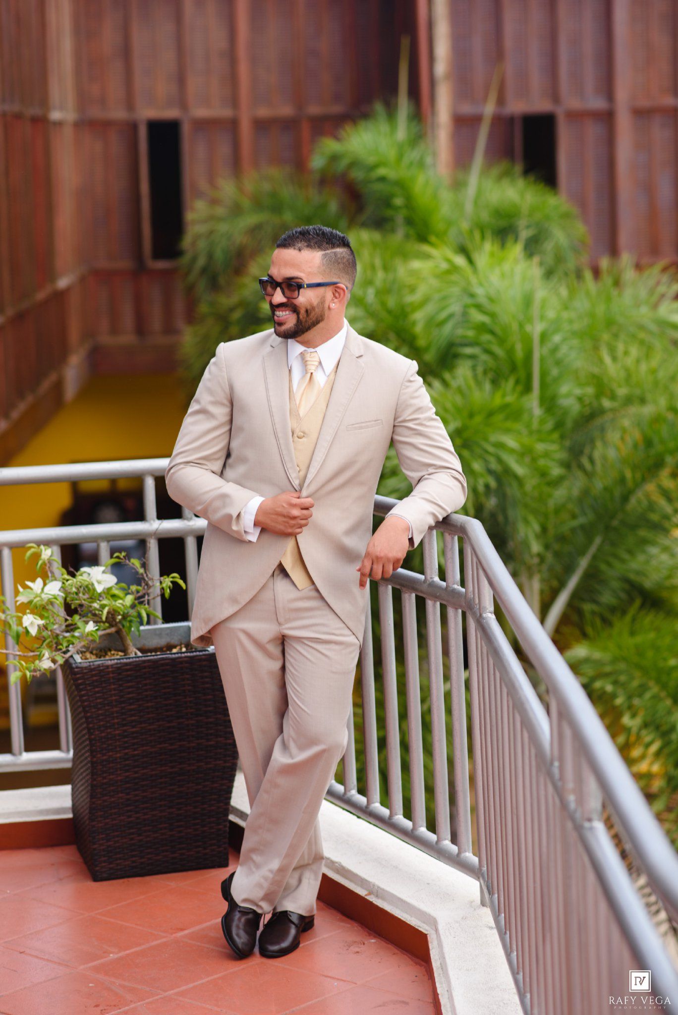 Groom in beige suit with gold vest and tie, wearing sunglasses, posing on outdoor terrace with lush greenery.
