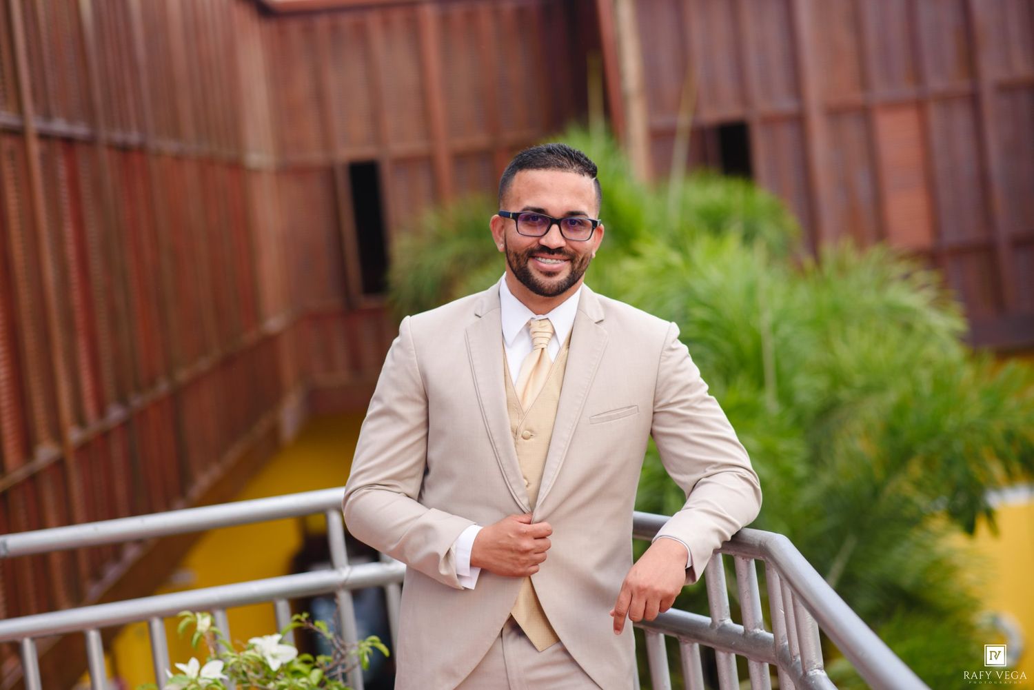Smiling man in beige suit and gold tie posing on outdoor walkway with greenery and rustic wall backdrop.