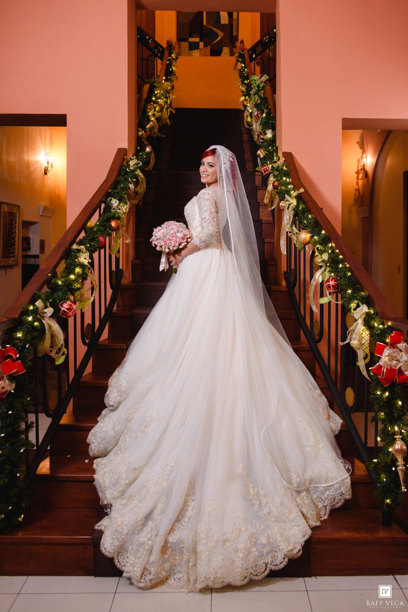 Bride in stunning lace ballgown with veil poses on staircase decorated with Christmas garlands and lights.