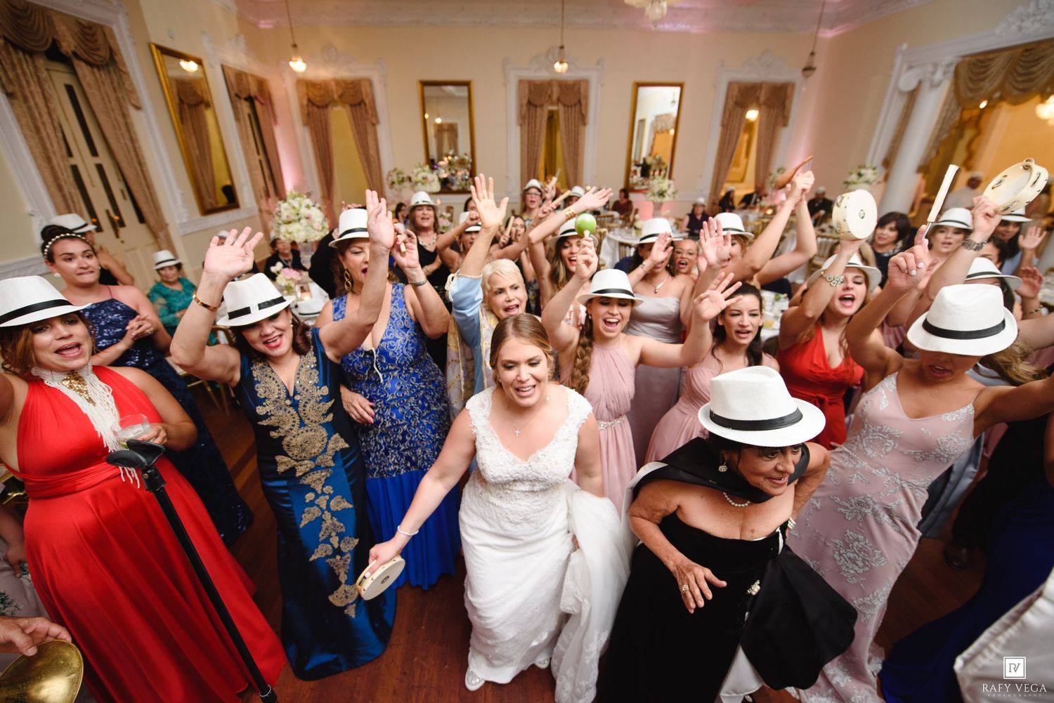 Bride surrounded by female wedding guests wearing white fedoras, celebrating joyfully on dance floor.