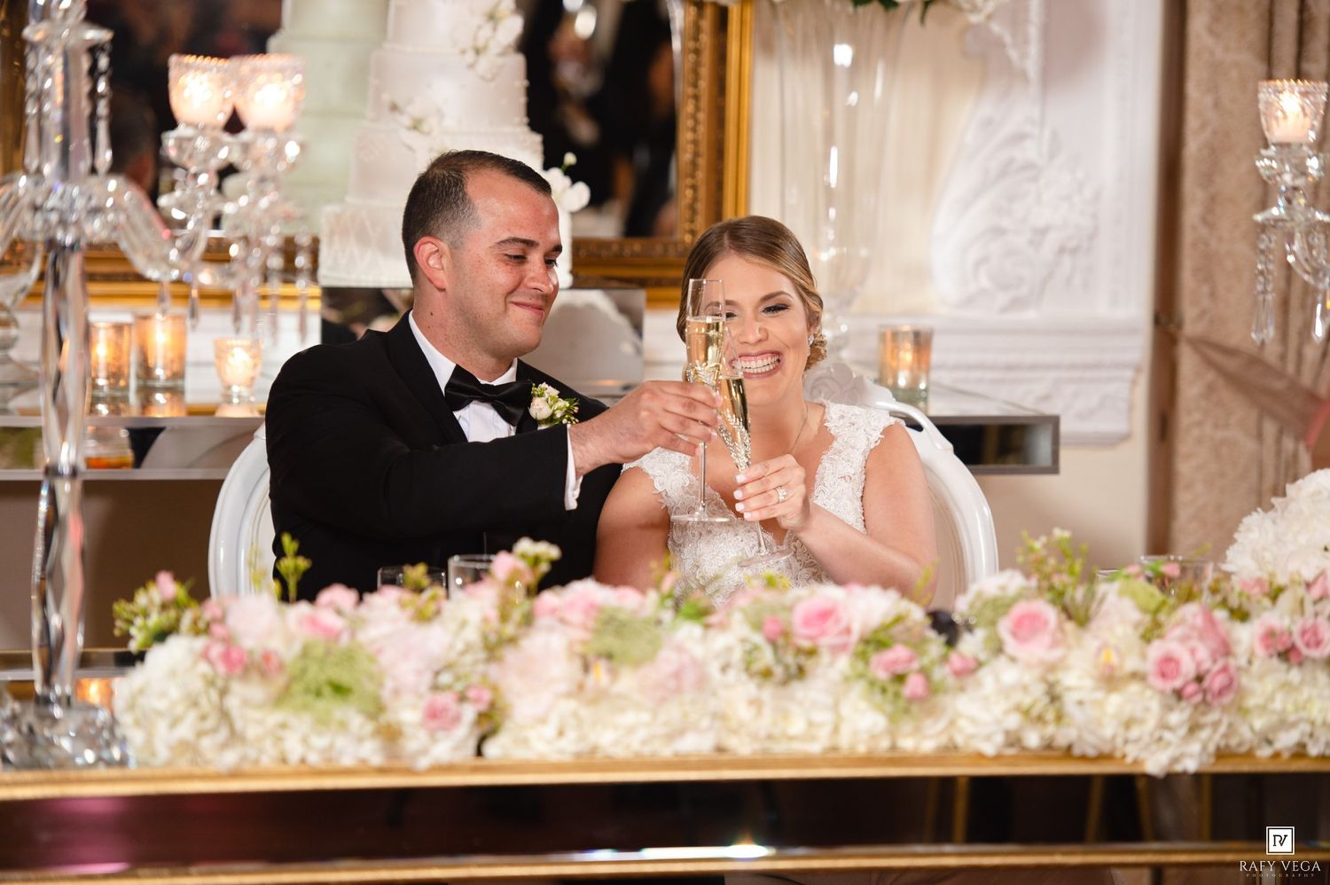 Bride and groom clink champagne glasses at sweetheart table during wedding reception toast.