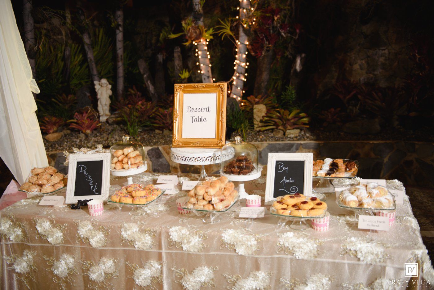 Elegant wedding dessert table with assorted pastries, chalkboard signs, and string lights at a reception.