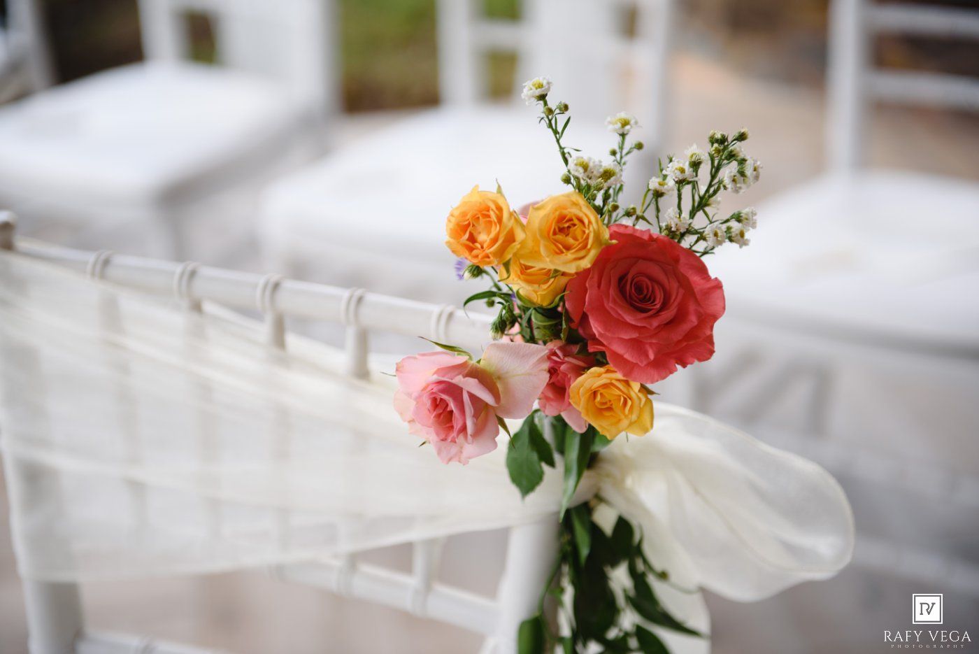 White chair adorned with colorful pink, orange, and yellow roses tied with sheer ribbon at a wedding ceremony.