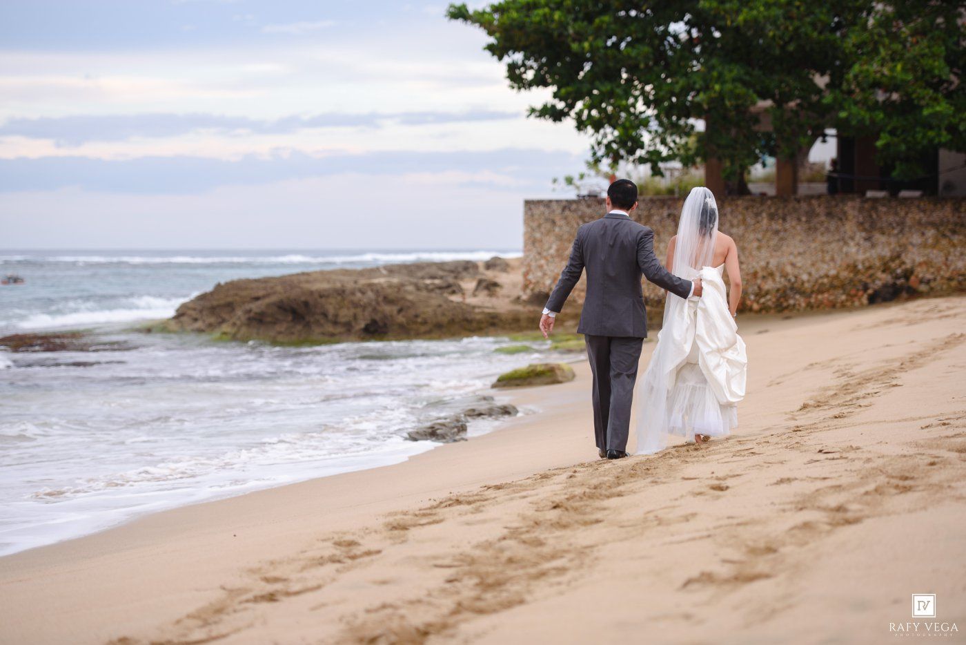 Bride and groom walking hand in hand along a sandy tropical beach, with ocean waves and lush greenery in the background.