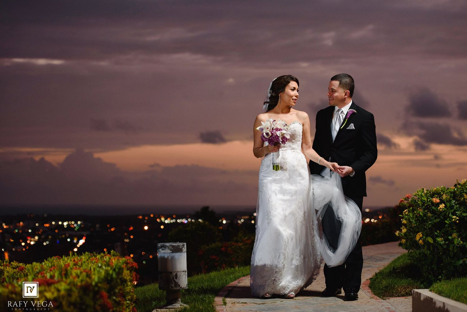 Bride and groom walking together at dusk with a stunning orange cloudy sky and city lights in the background.