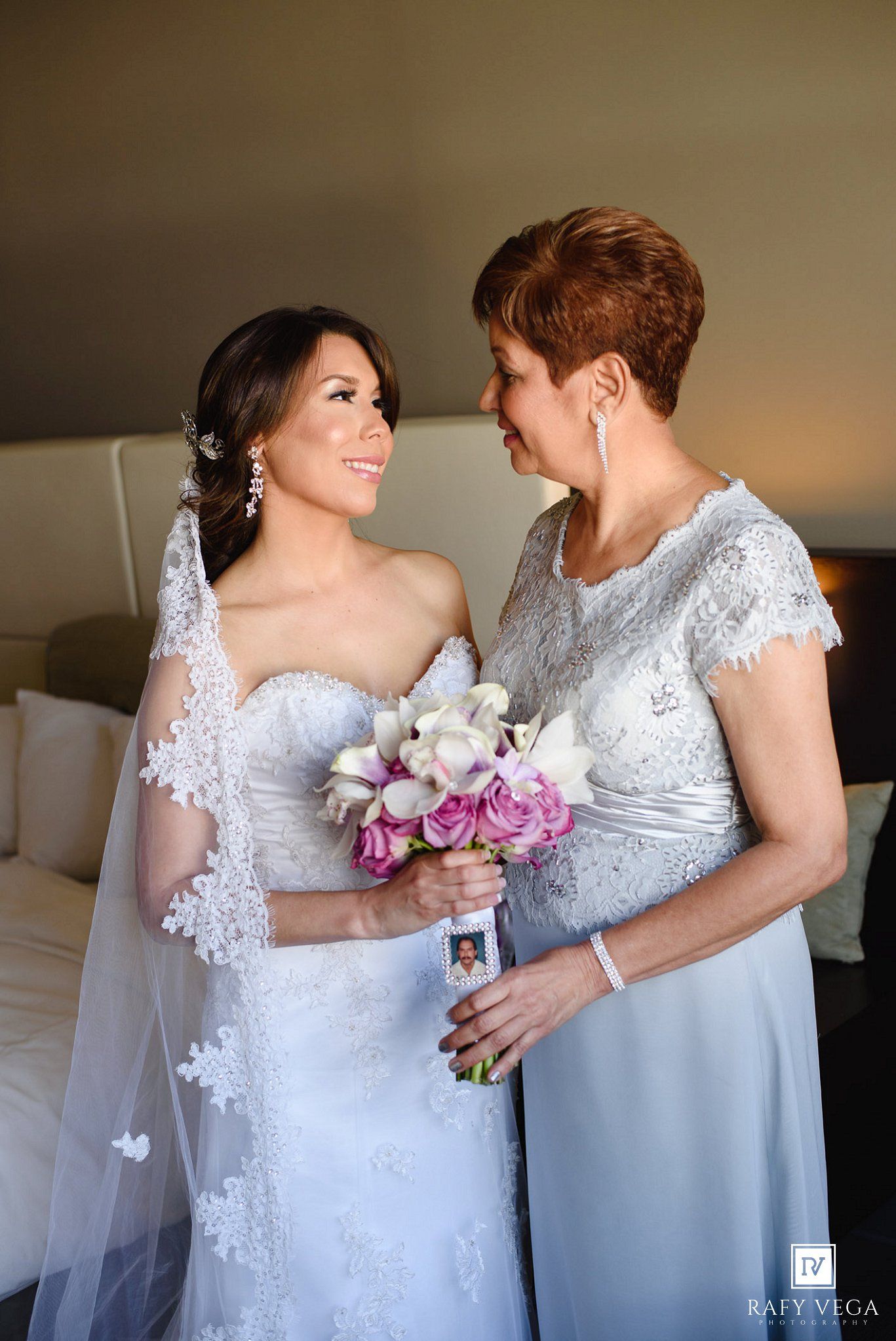 Bride in white lace gown holding floral bouquet smiles at older woman in silver dress during wedding preparation.