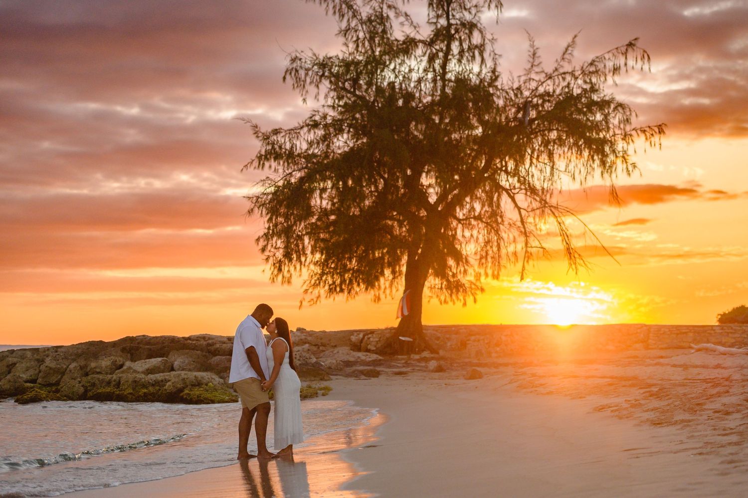 Couple sharing a romantic moment on a tropical beach at golden sunset with a silhouetted tree backdrop.