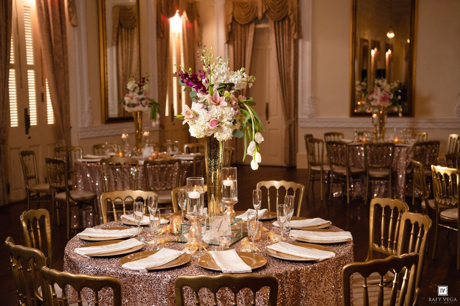 Luxurious gold sequin tablecloth with tall floral centerpiece of white and purple blooms at wedding reception.