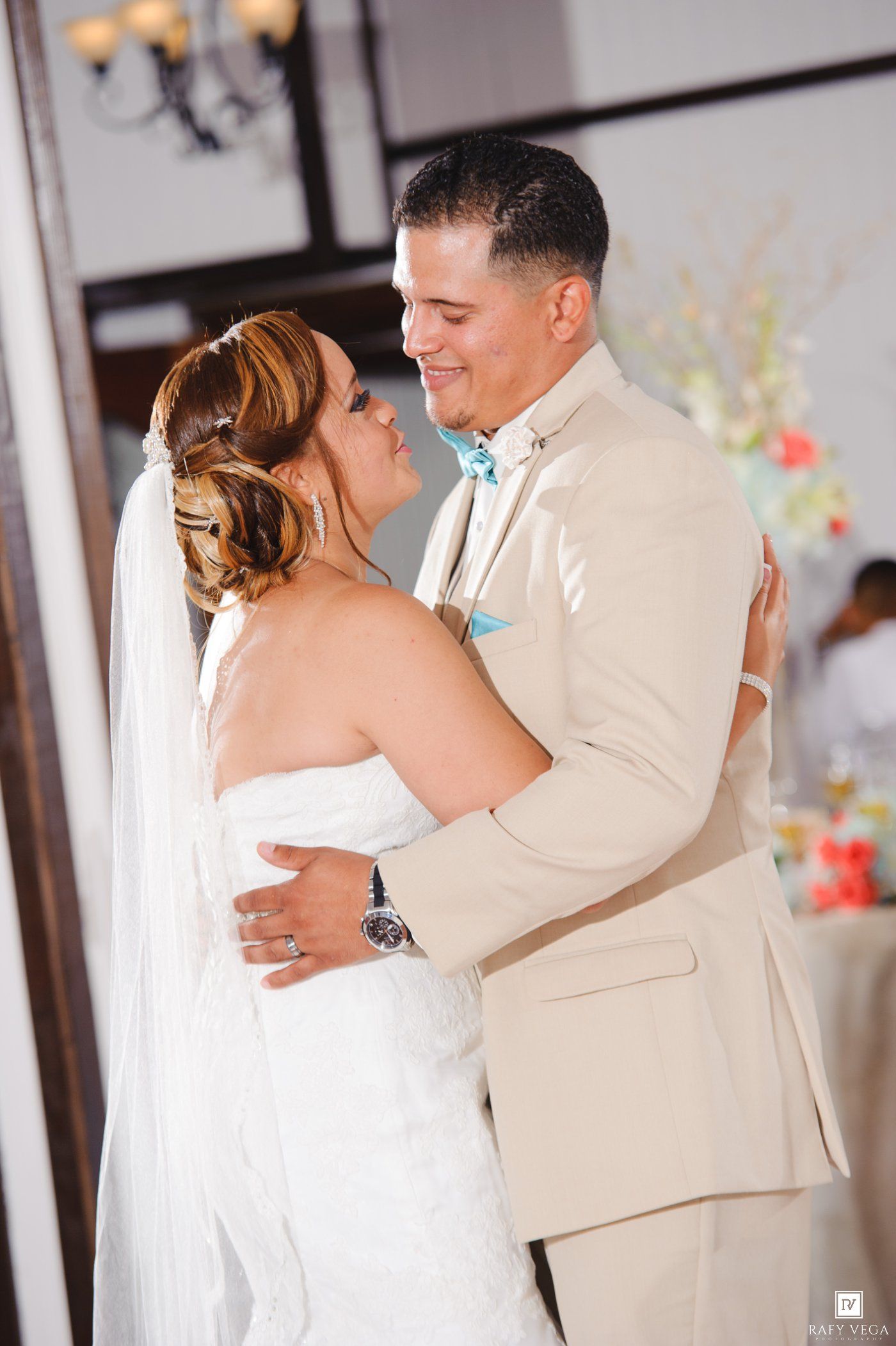 Bride and groom share their first dance, she in a white strapless gown with veil, he in a beige suit with teal bow tie.