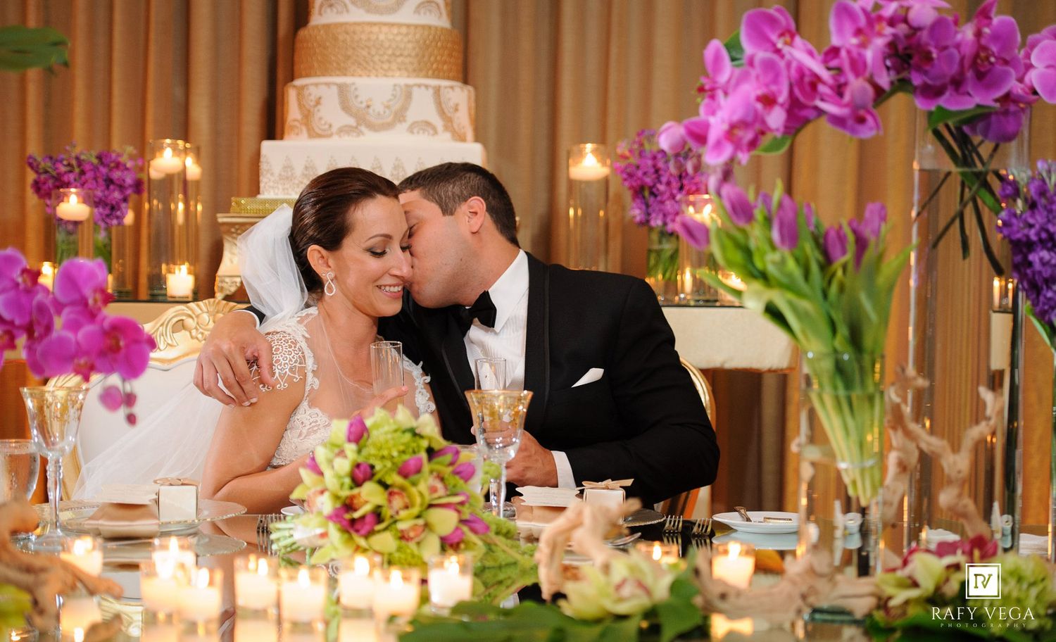Bride and groom share a romantic kiss at wedding reception sweetheart table surrounded by candles and purple orchid floral arrangements.