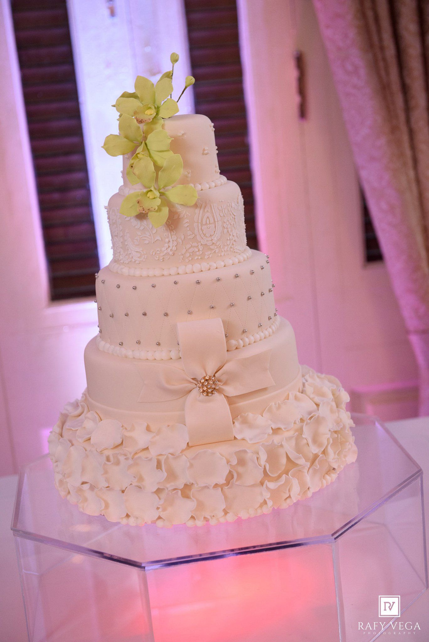 Elegant four-tier white wedding cake with green orchids, pearl details, bow, and ruffles on illuminated stand.
