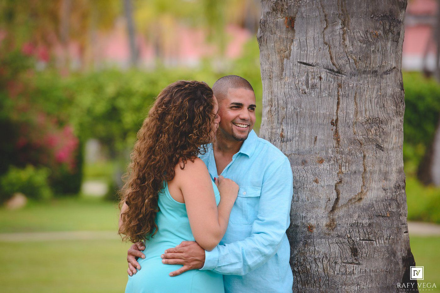Smiling couple embracing by a palm tree in a lush tropical garden, wearing matching turquoise outfits.