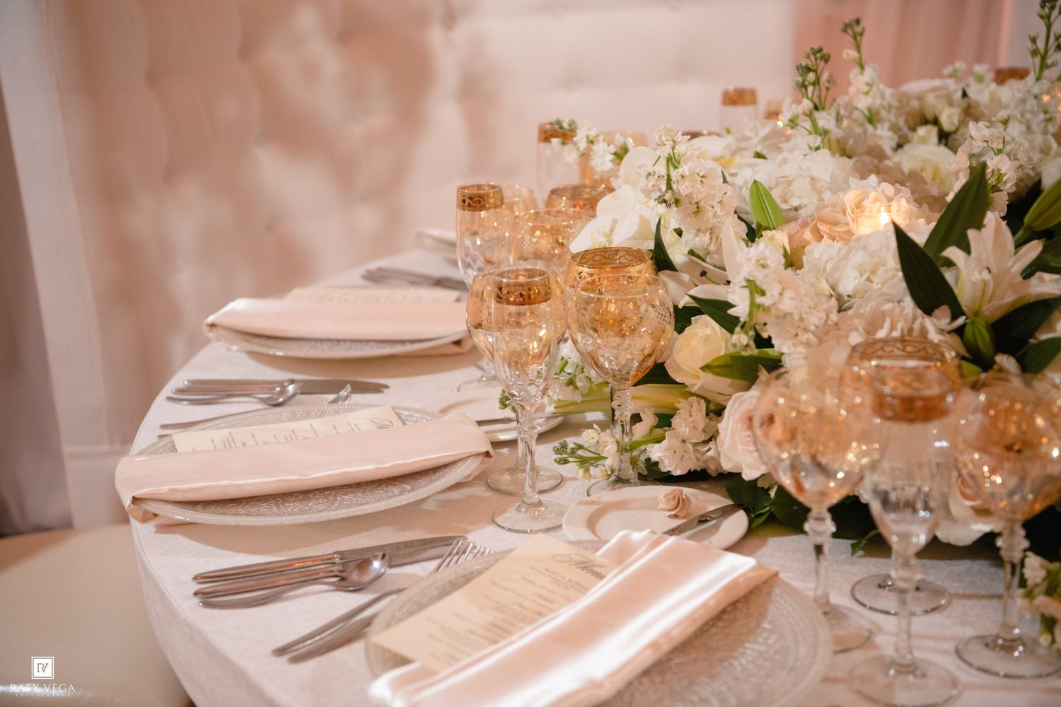 Elegant wedding reception table setting with gold glassware, white floral centerpiece, and silver cutlery.