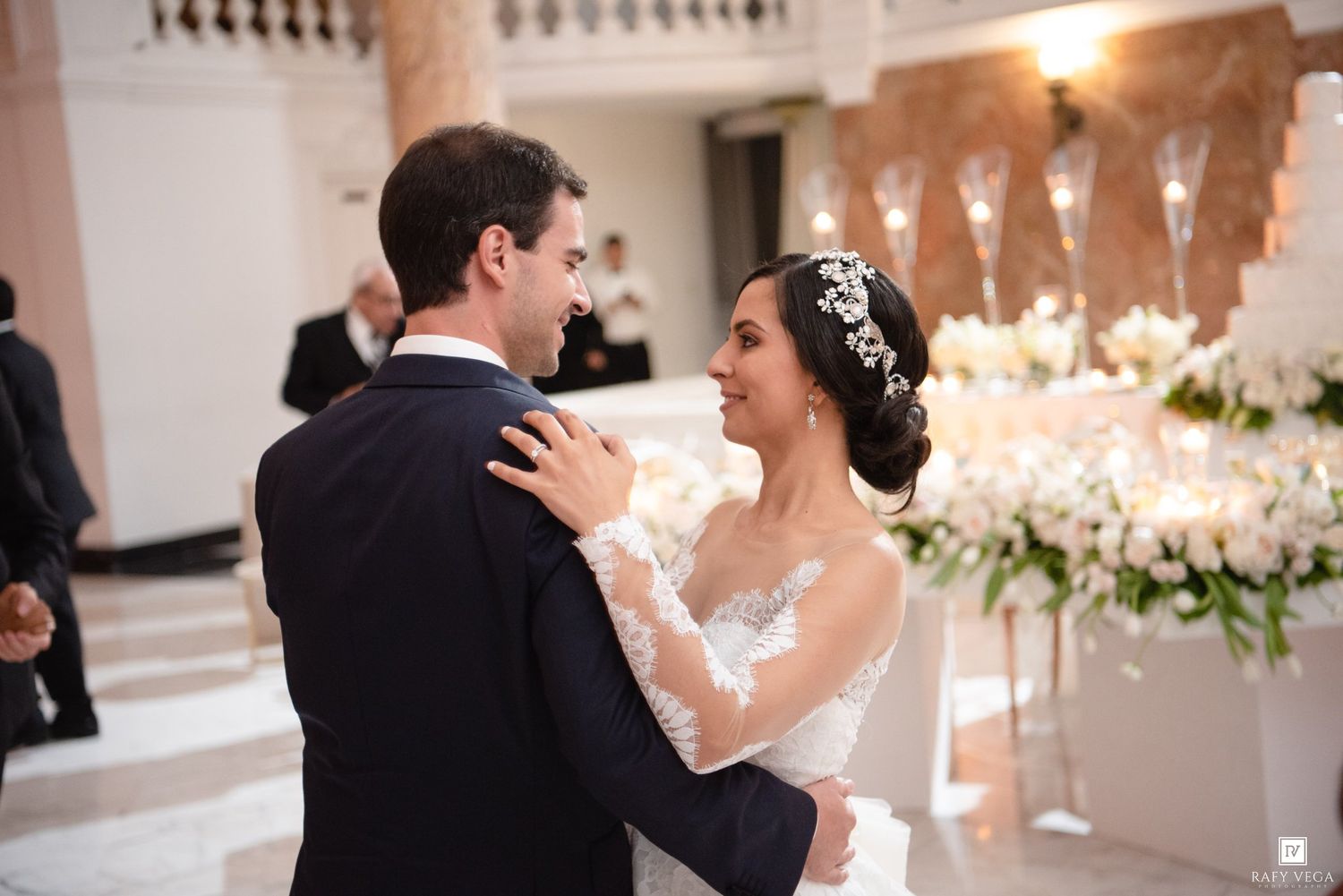 Bride and groom share first dance in elegant wedding venue with white floral arrangements.