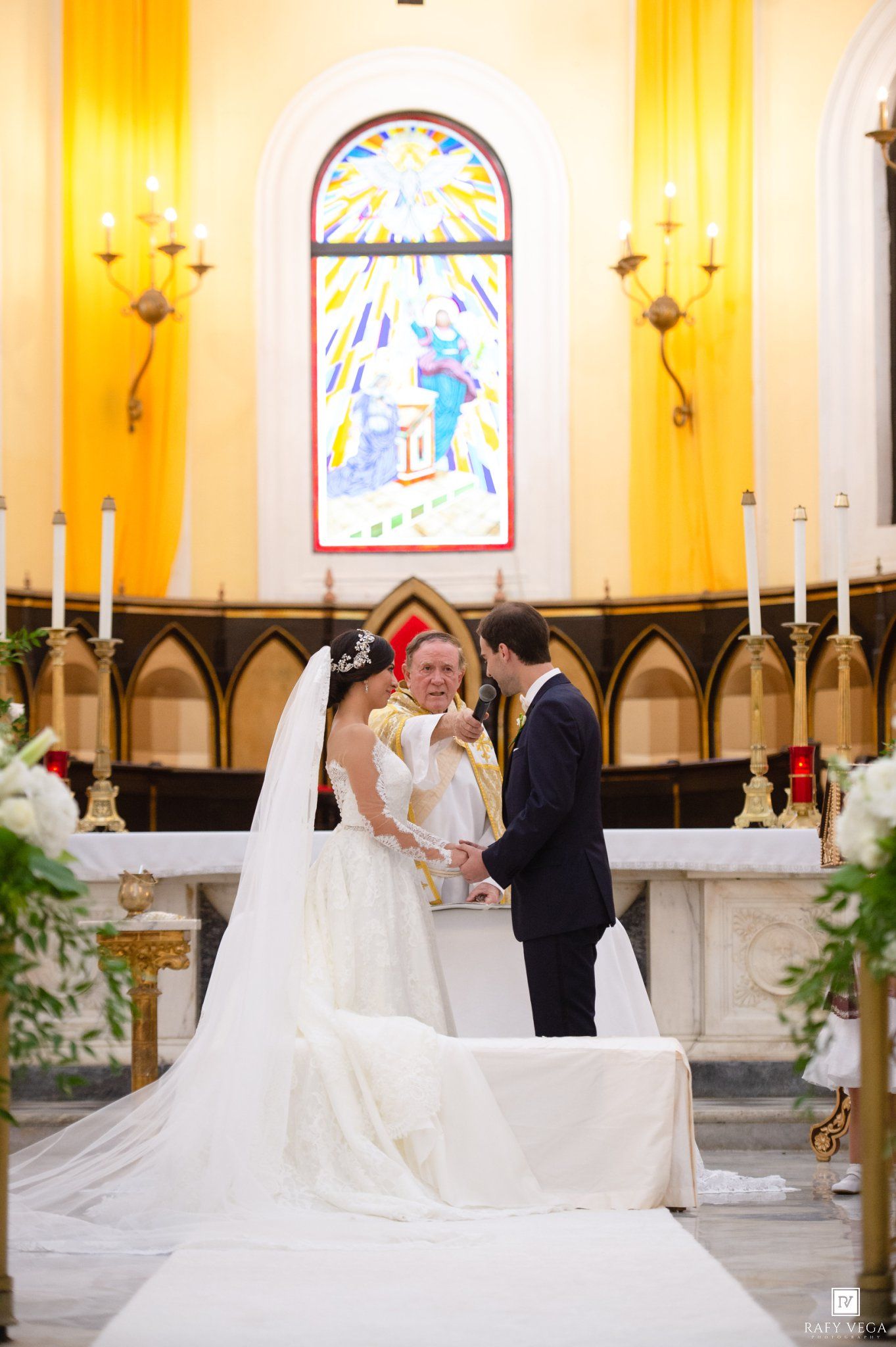 Bride and groom exchanging vows at church altar with colorful stained glass window and candles behind them.