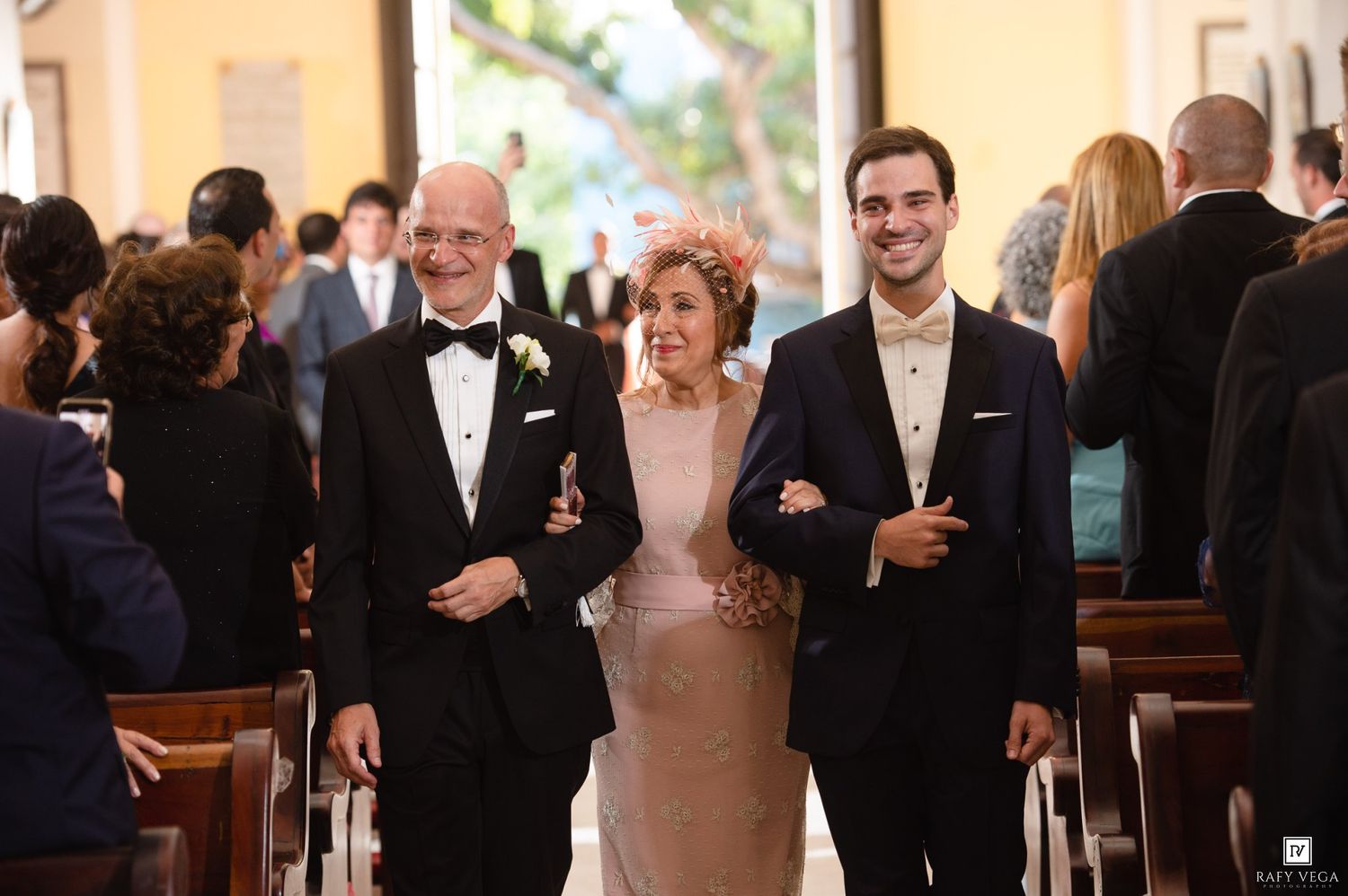A smiling family of three walks down a church aisle at a wedding, dressed in formal attire including tuxedos and an elegant gown.