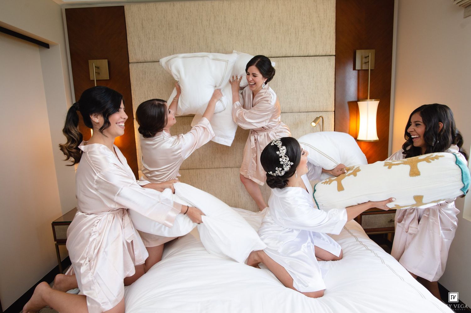 Bridesmaids in pink satin robes having a fun pillow fight on a hotel bed before the wedding ceremony.