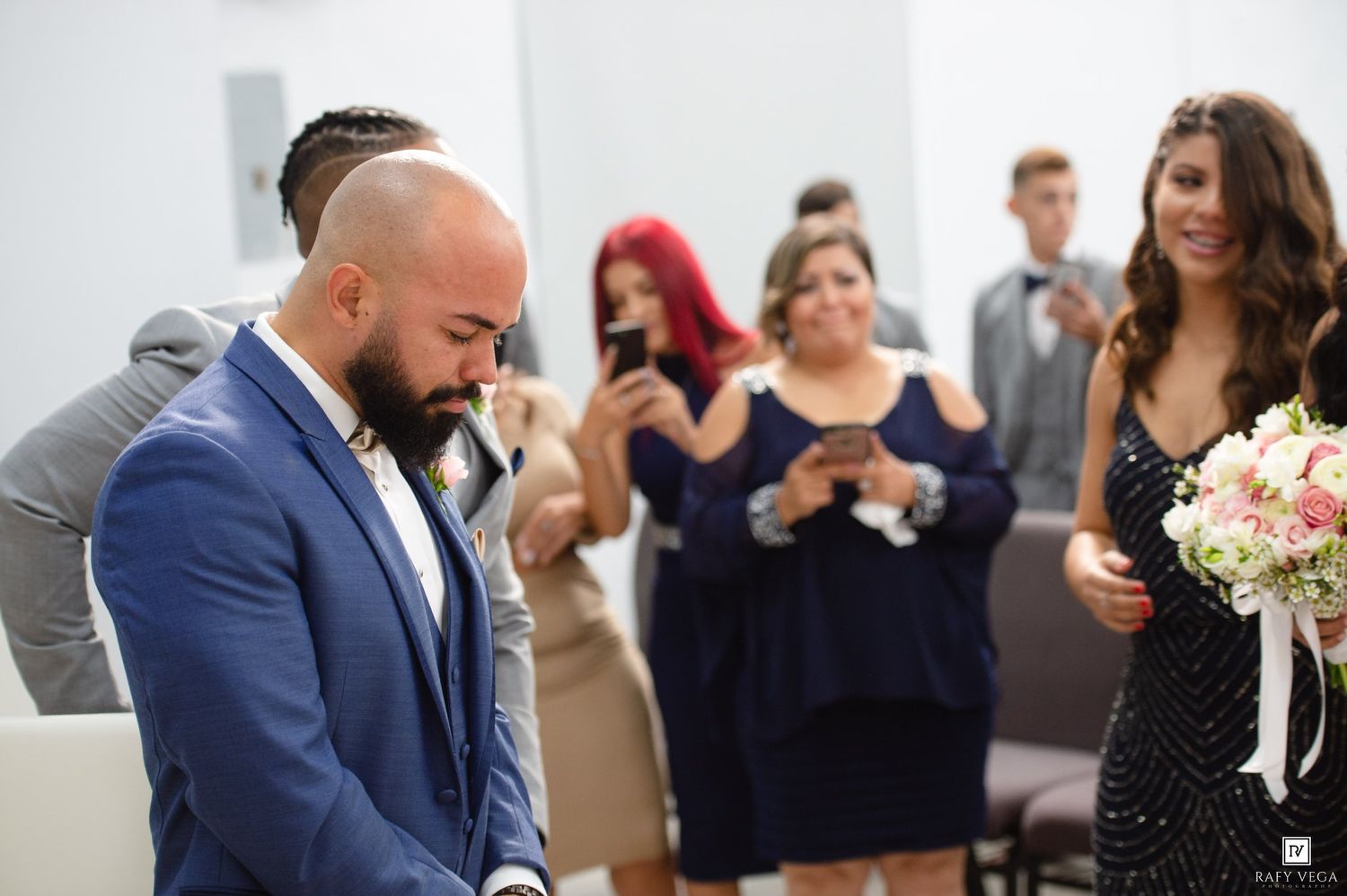 Emotional groom looks down during wedding ceremony as guests capture the moment on phones in elegant venue.