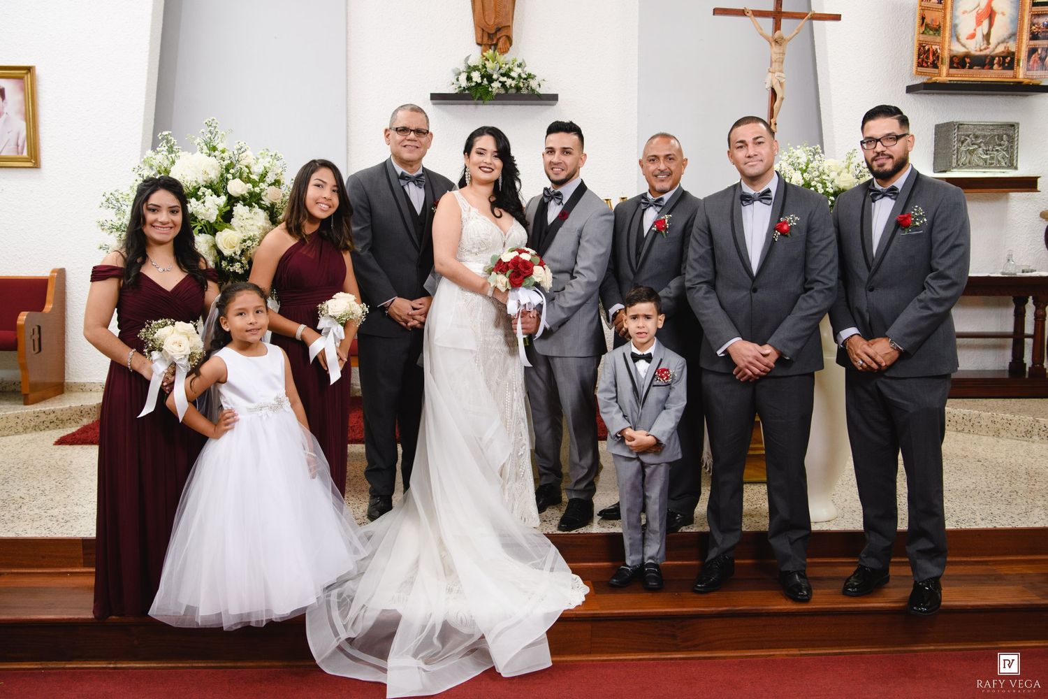 Wedding party posing in church, bride in white gown, groom in gray suit, bridesmaids in burgundy dresses.