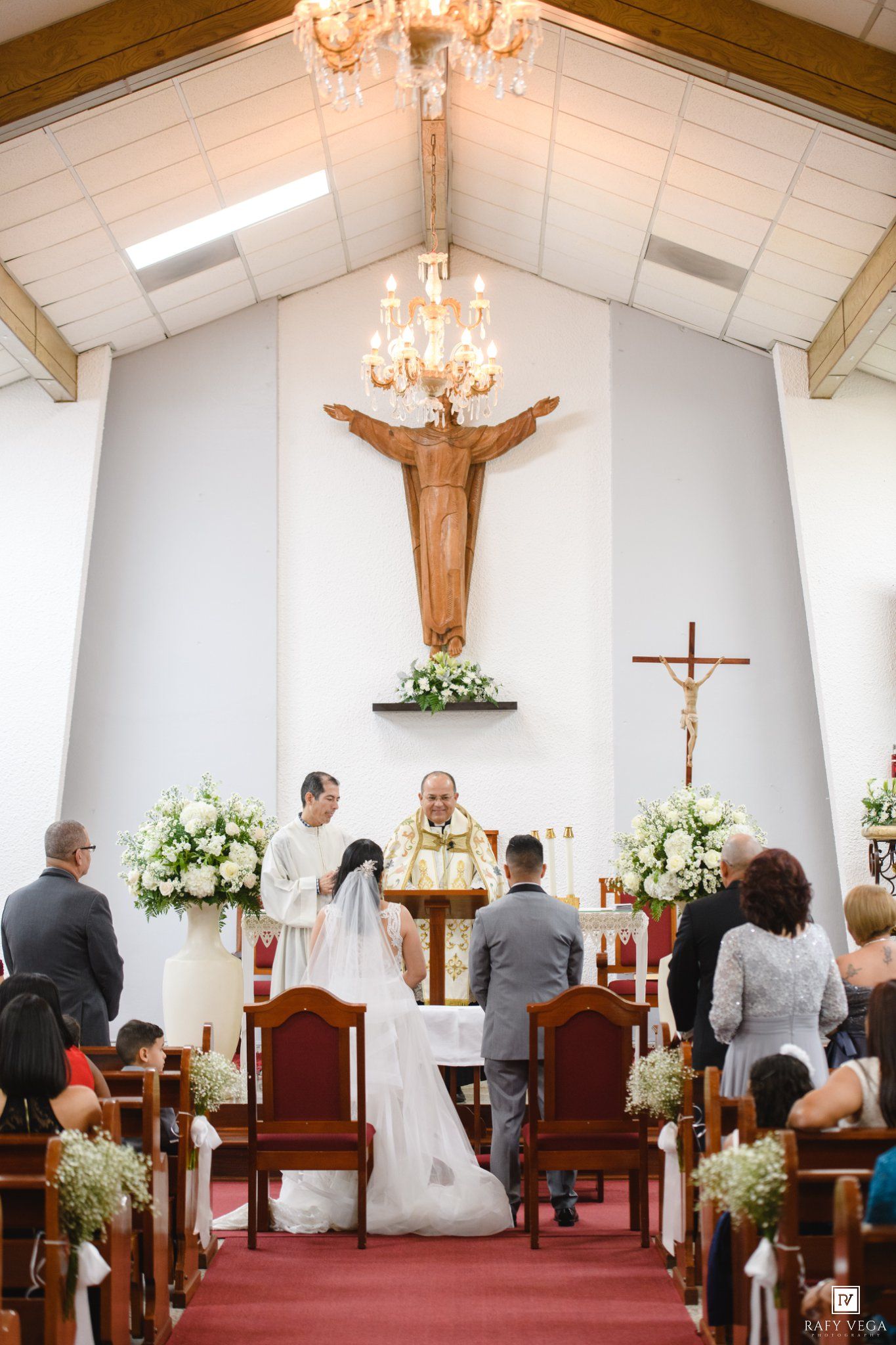 A bride and groom stand at the altar during a Catholic wedding ceremony in a white chapel with a wooden crucifix.