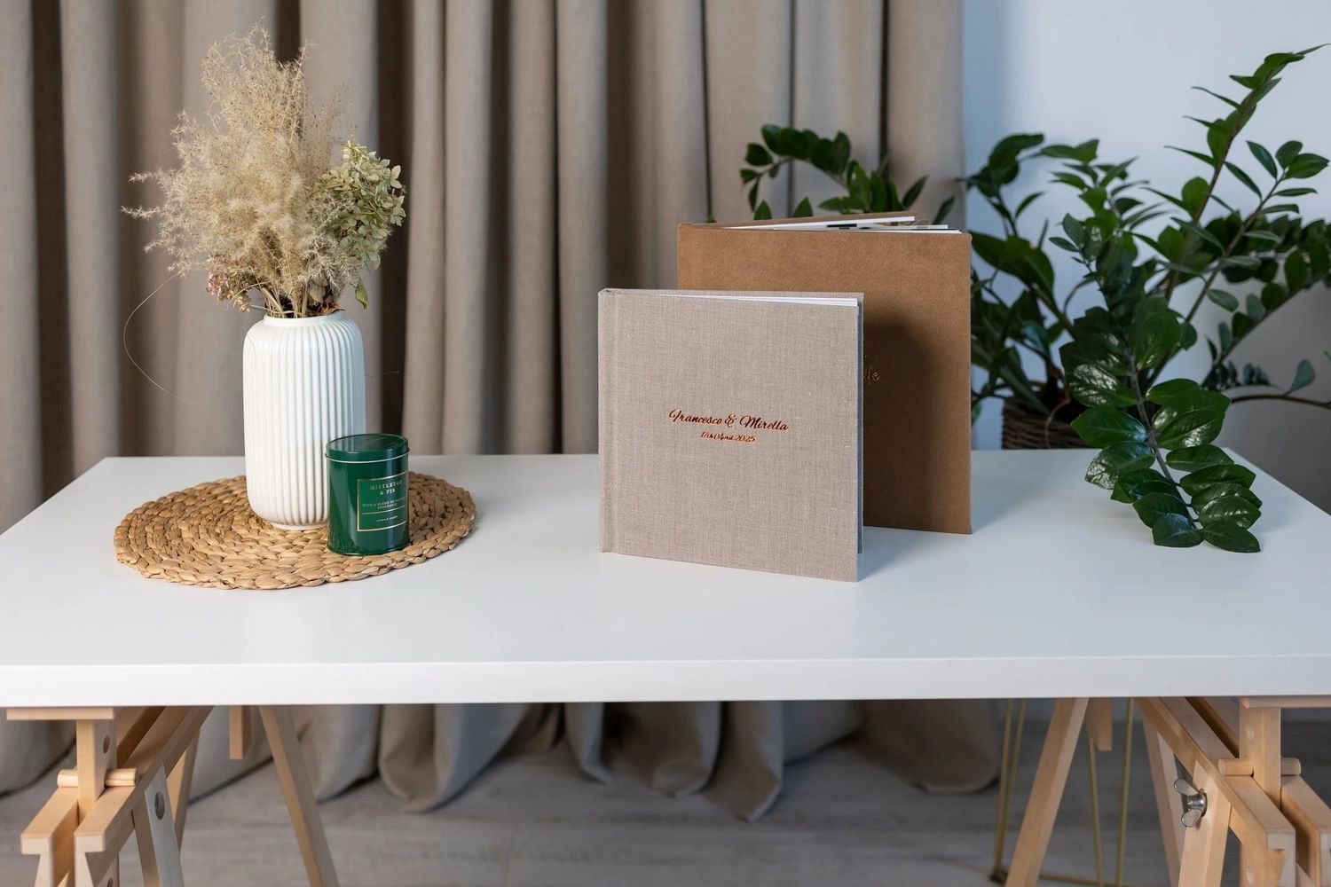 Elegant linen photo album on a white trestle table with a vase, green candle, and plants in a neutral setting.