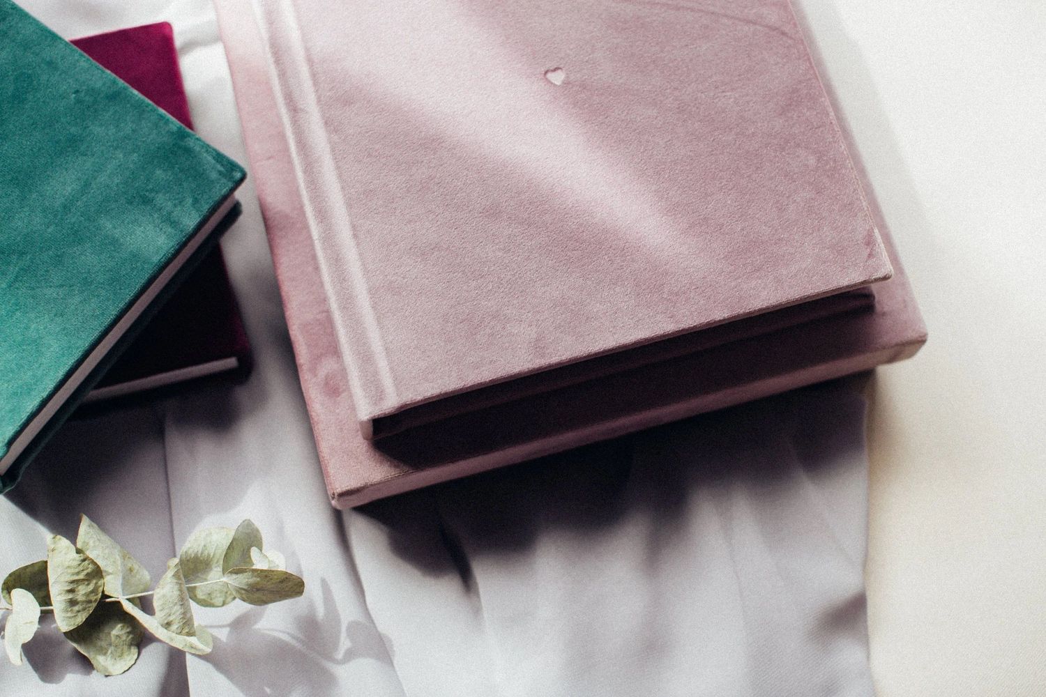 Pink and teal hardcover books stacked on white fabric with eucalyptus leaves, elegant flat lay photography.
