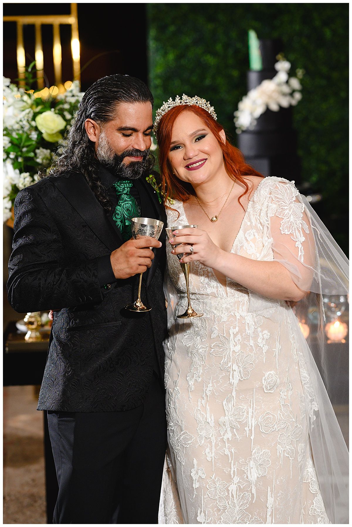 Bride in lace gown and groom in black suit share a toast with elegant glasses at their wedding reception.