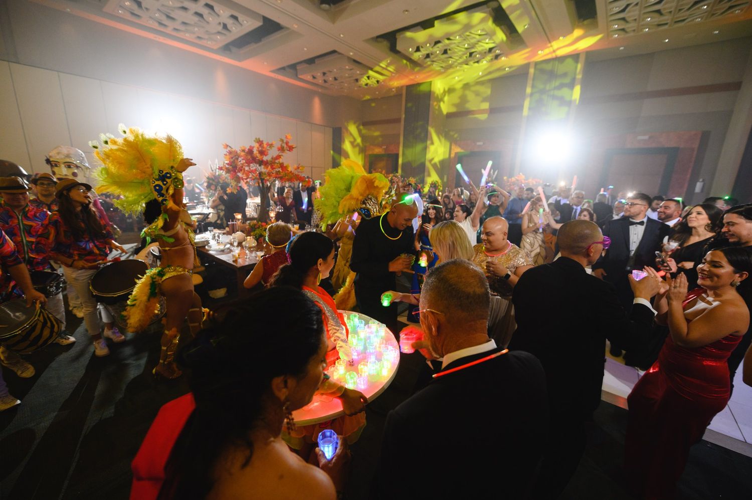 Festive gala event with guests in formal attire dancing alongside colorful feathered performers under dramatic stage lighting.