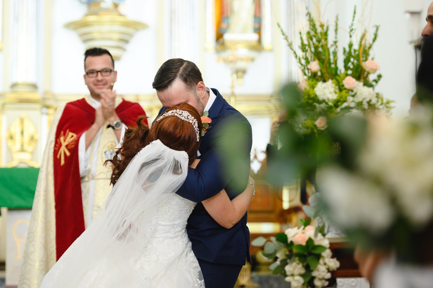 Bride and groom share first kiss at church altar with priest in red vestments during wedding ceremony.