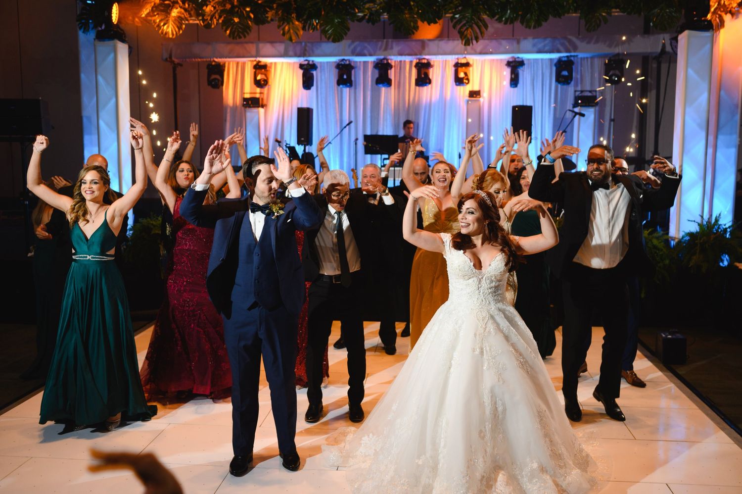 Bride and groom celebrate with wedding party on dance floor, arms raised in joy at elegant reception venue.