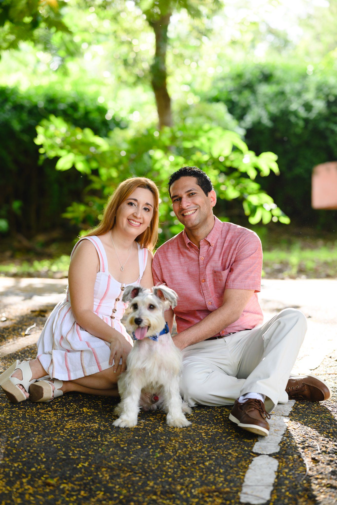 Happy couple sitting on ground with small white Schnauzer dog in lush green park setting.