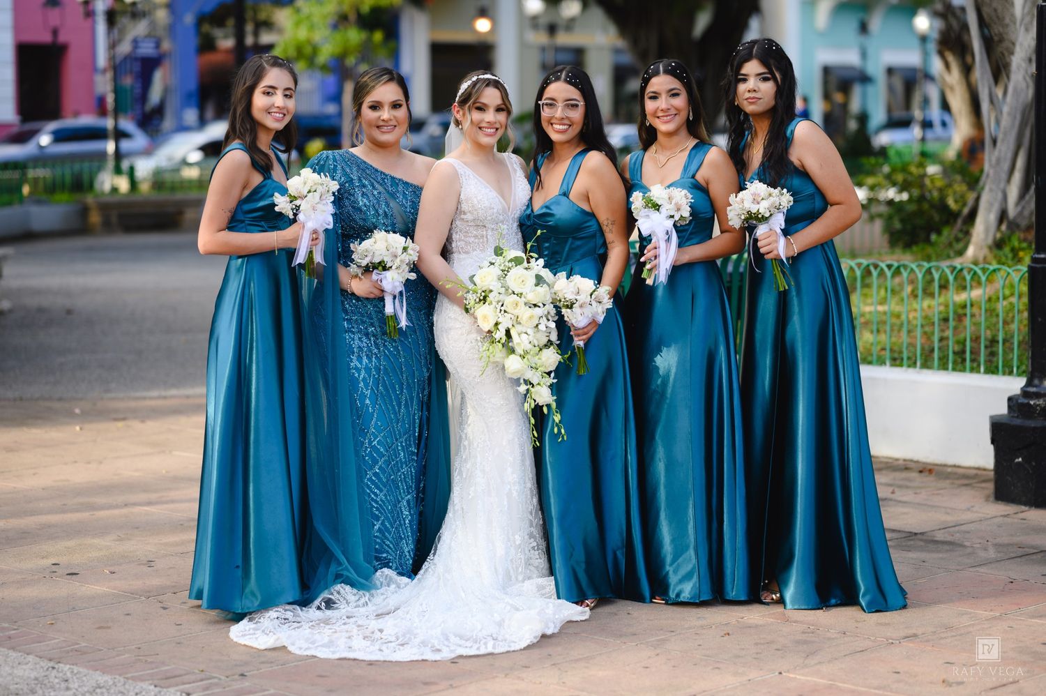 Bride in white lace gown poses with five bridesmaids in teal satin dresses holding bouquets outdoors.