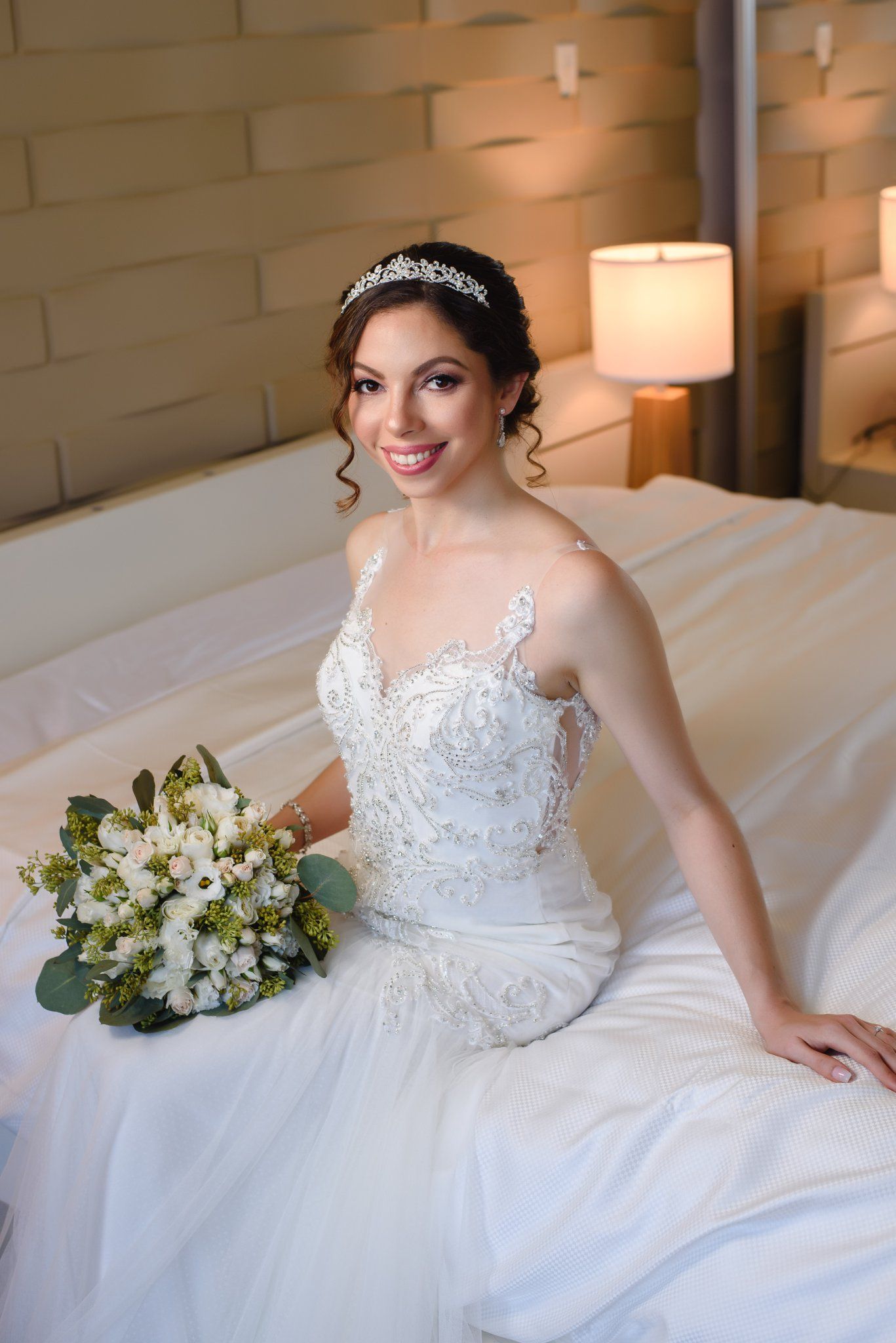 Beautiful bride in white lace gown sitting on bed holding white floral bouquet, wearing silver tiara.