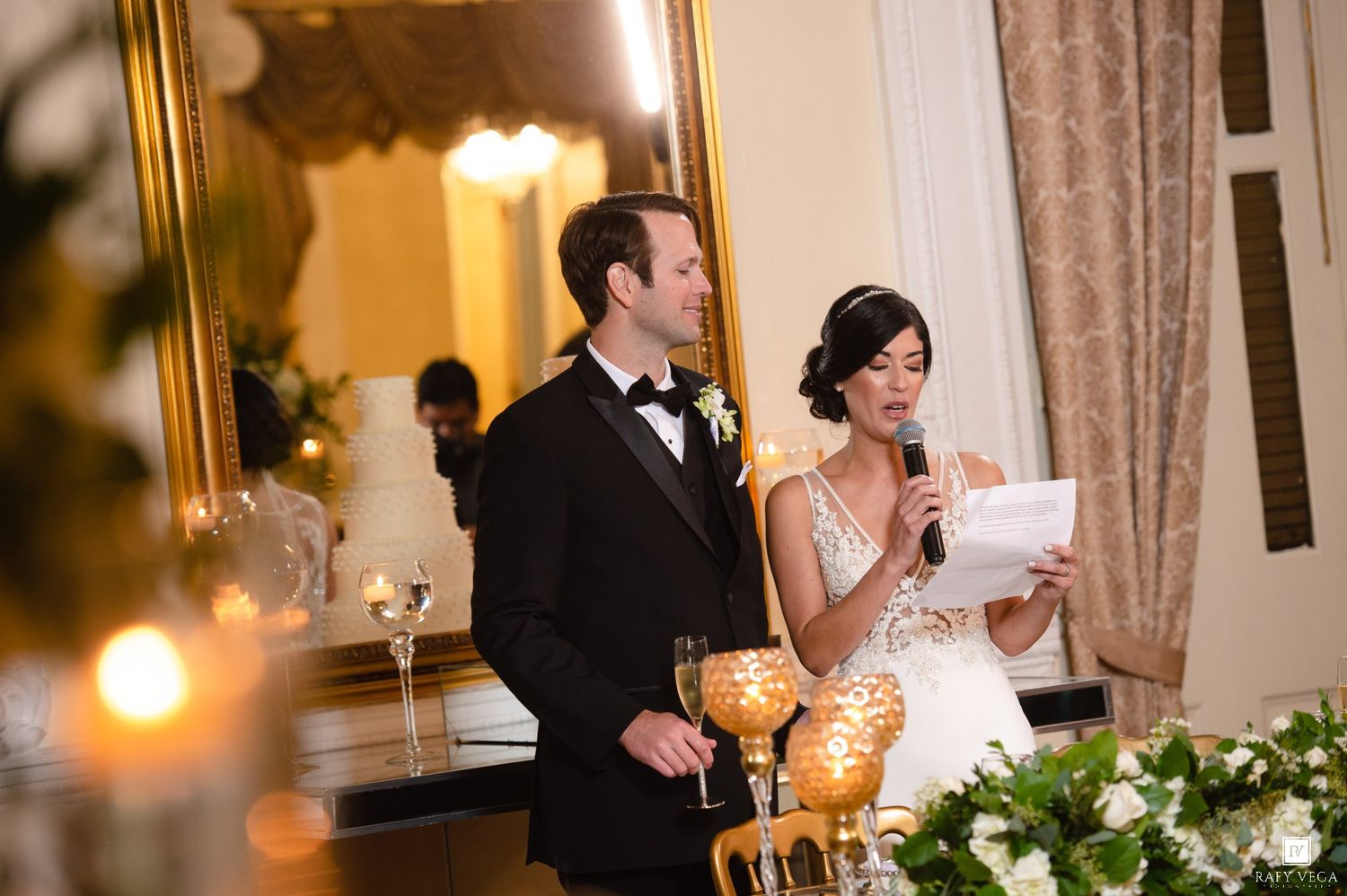 Bride reads speech into microphone while groom stands beside her at elegant wedding reception.