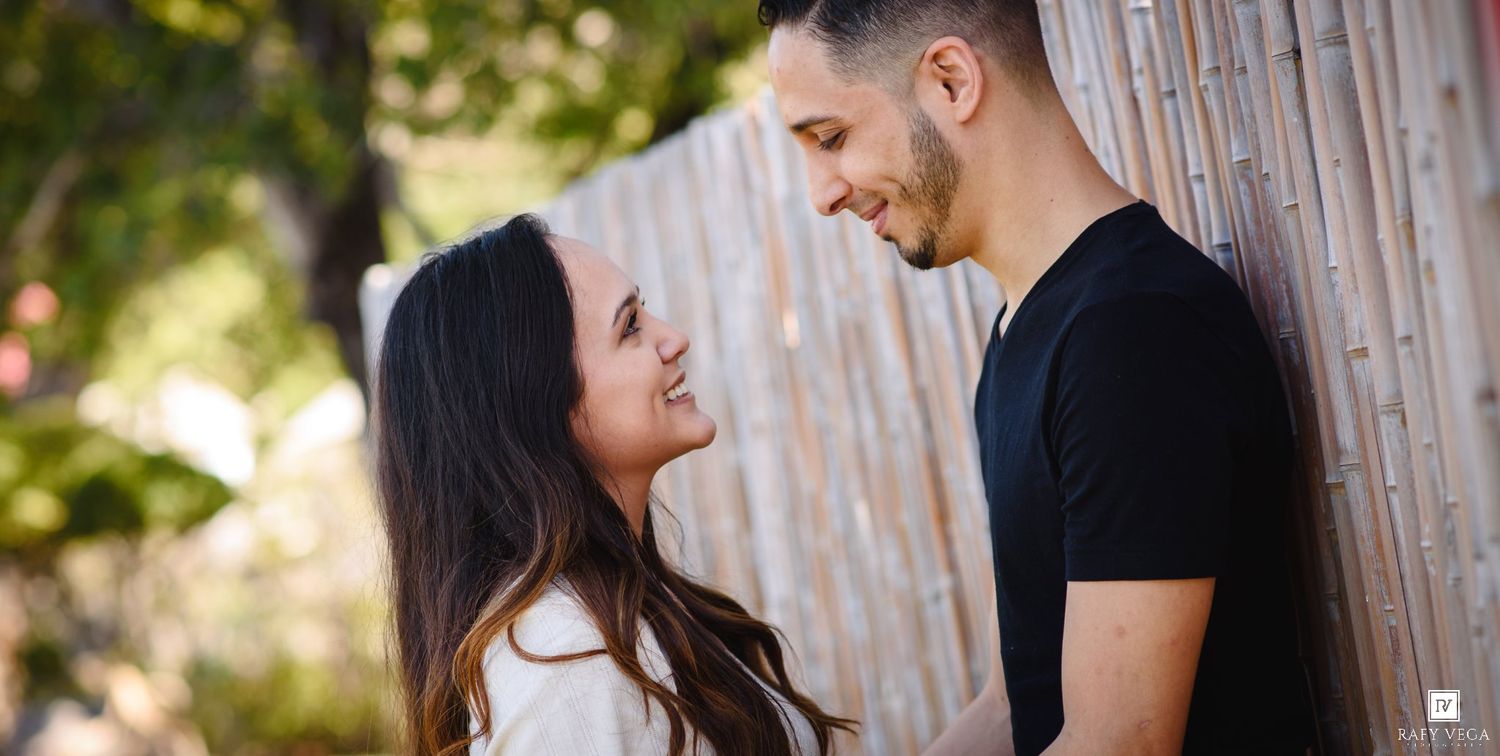 A smiling couple sharing a romantic moment outdoors near a wooden fence surrounded by greenery.