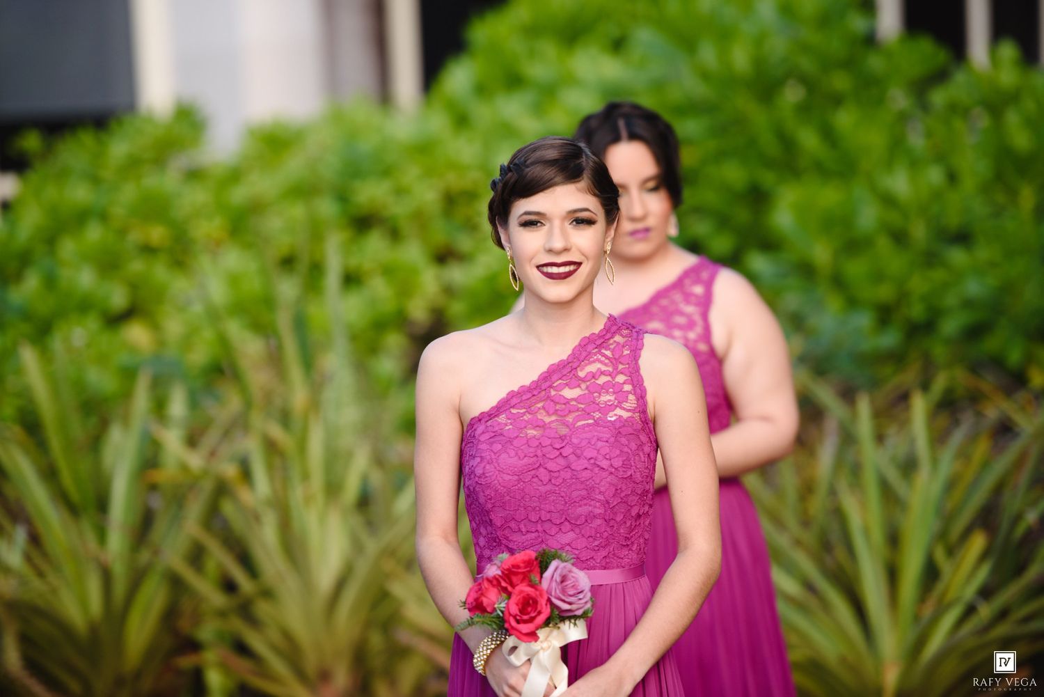 Two bridesmaids in purple lace one-shoulder gowns holding bouquets pose outdoors at wedding.